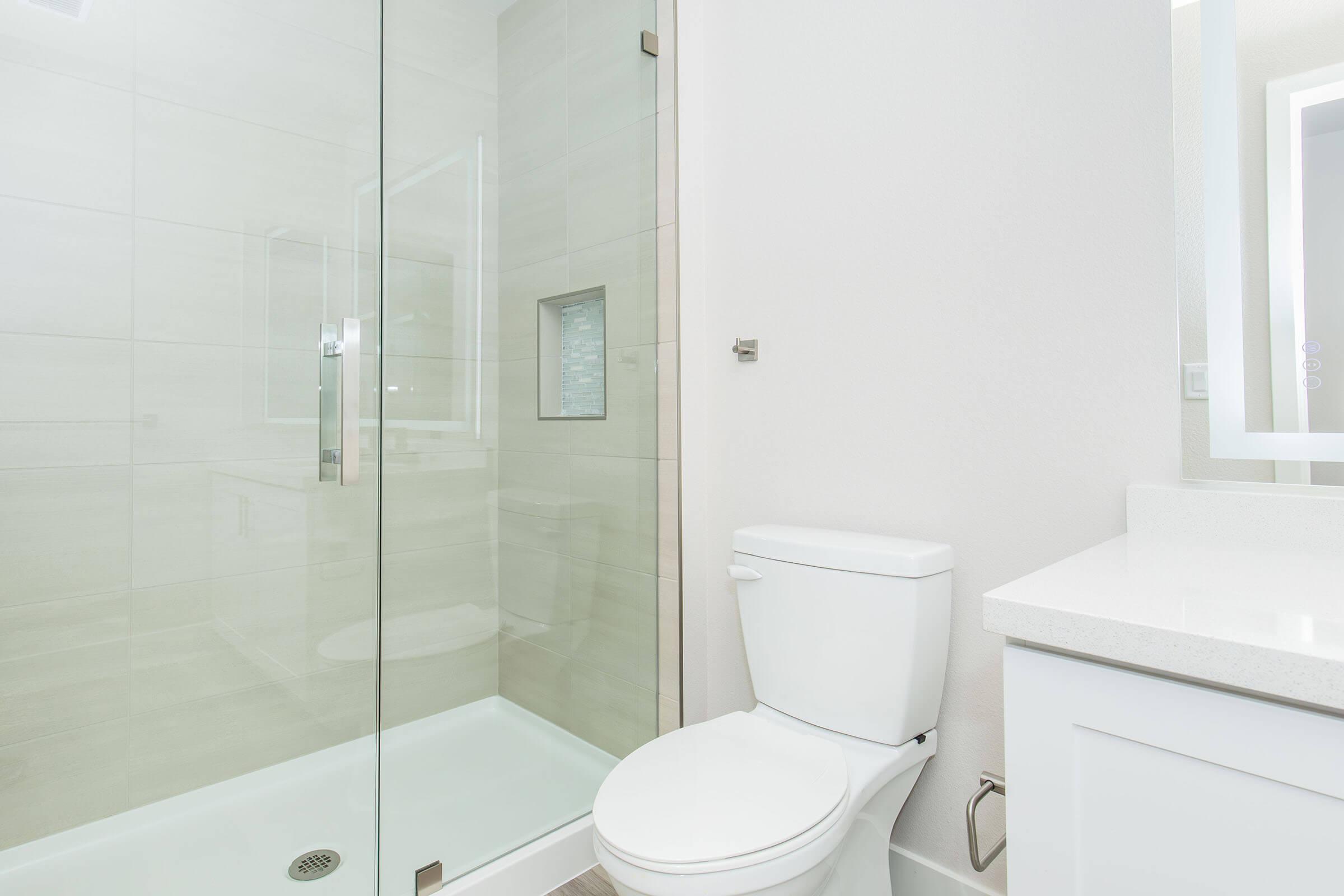 A modern bathroom featuring a glass shower enclosure, a white toilet, and a sleek vanity with a mirror. The walls are tiled in light colors, contributing to a clean and spacious appearance. Natural light enters the room, enhancing the bright ambiance.