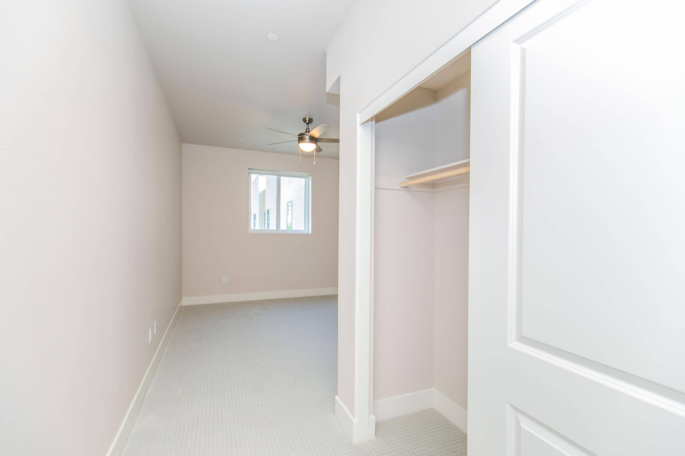 A simple, empty room featuring light-colored walls and carpet. There is a window allowing natural light, a ceiling fan, and an open closet with a shelf. The space is uncluttered, creating a bright and airy atmosphere, suitable for various uses such as a bedroom or office.