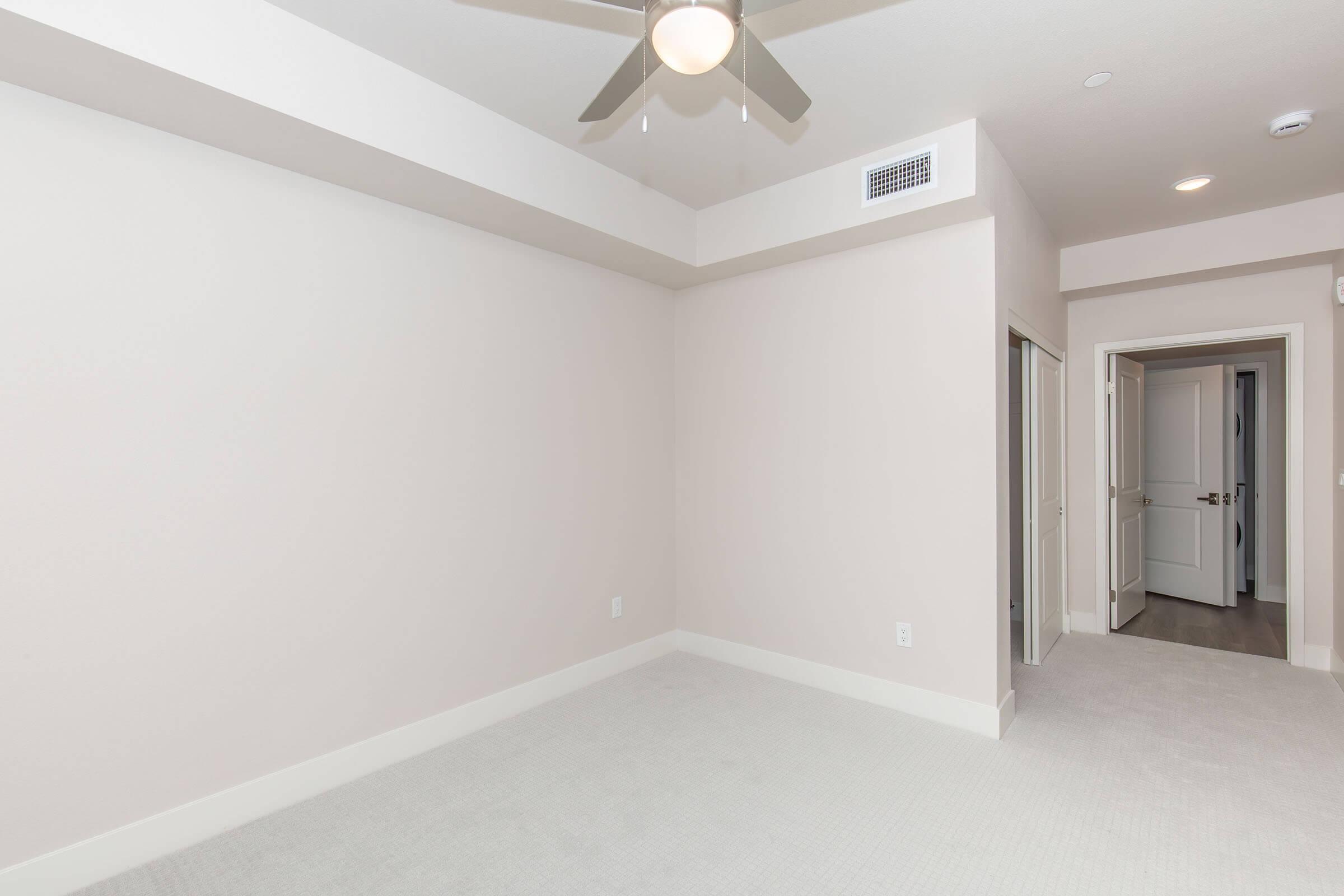 Empty room with light-colored walls and carpeted floor. A ceiling fan is installed in the center, and there are two doorways leading to other areas. The room has a bright and neutral aesthetic, with no furniture present.