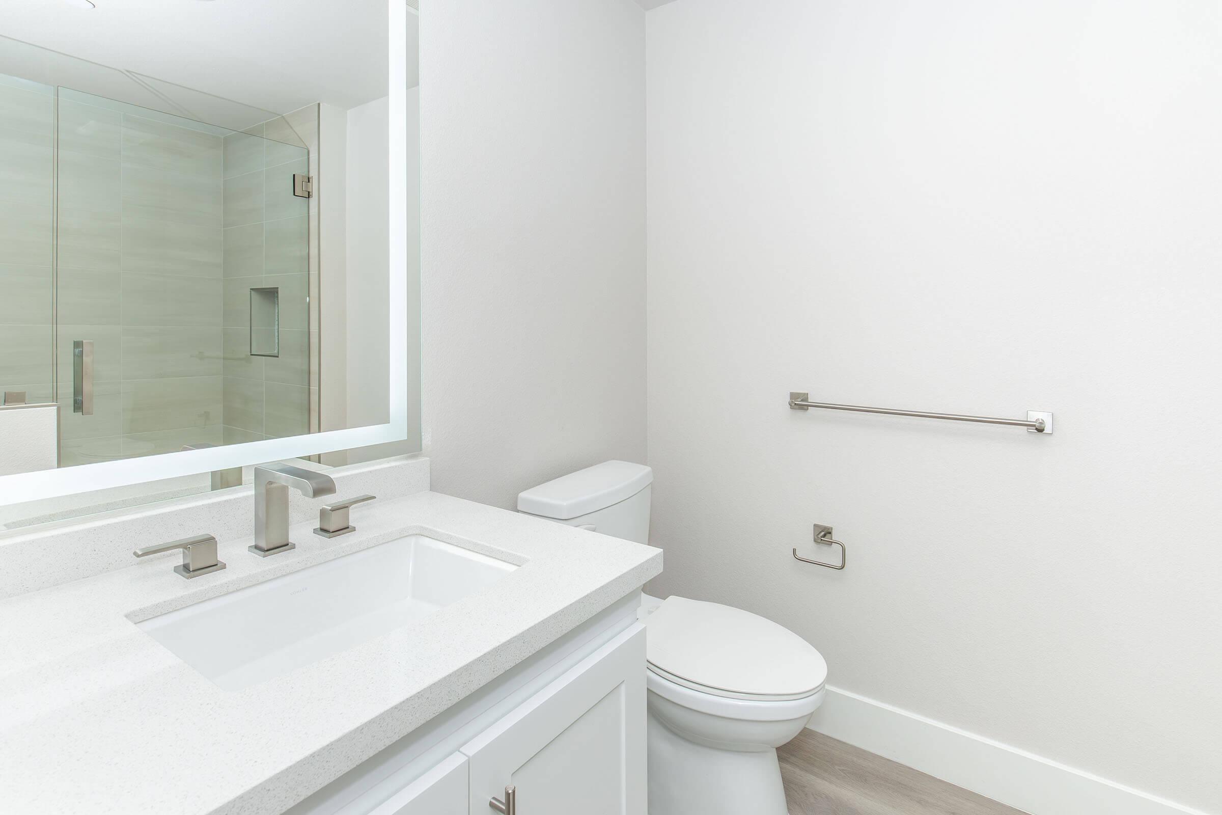 Modern bathroom featuring a clean design with a large mirror above a white sink with sleek faucets. A white toilet is positioned beside the sink, with a wall-mounted towel bar nearby. The space has neutral tones and a light, airy feel, complemented by a stylish shower area behind glass doors.