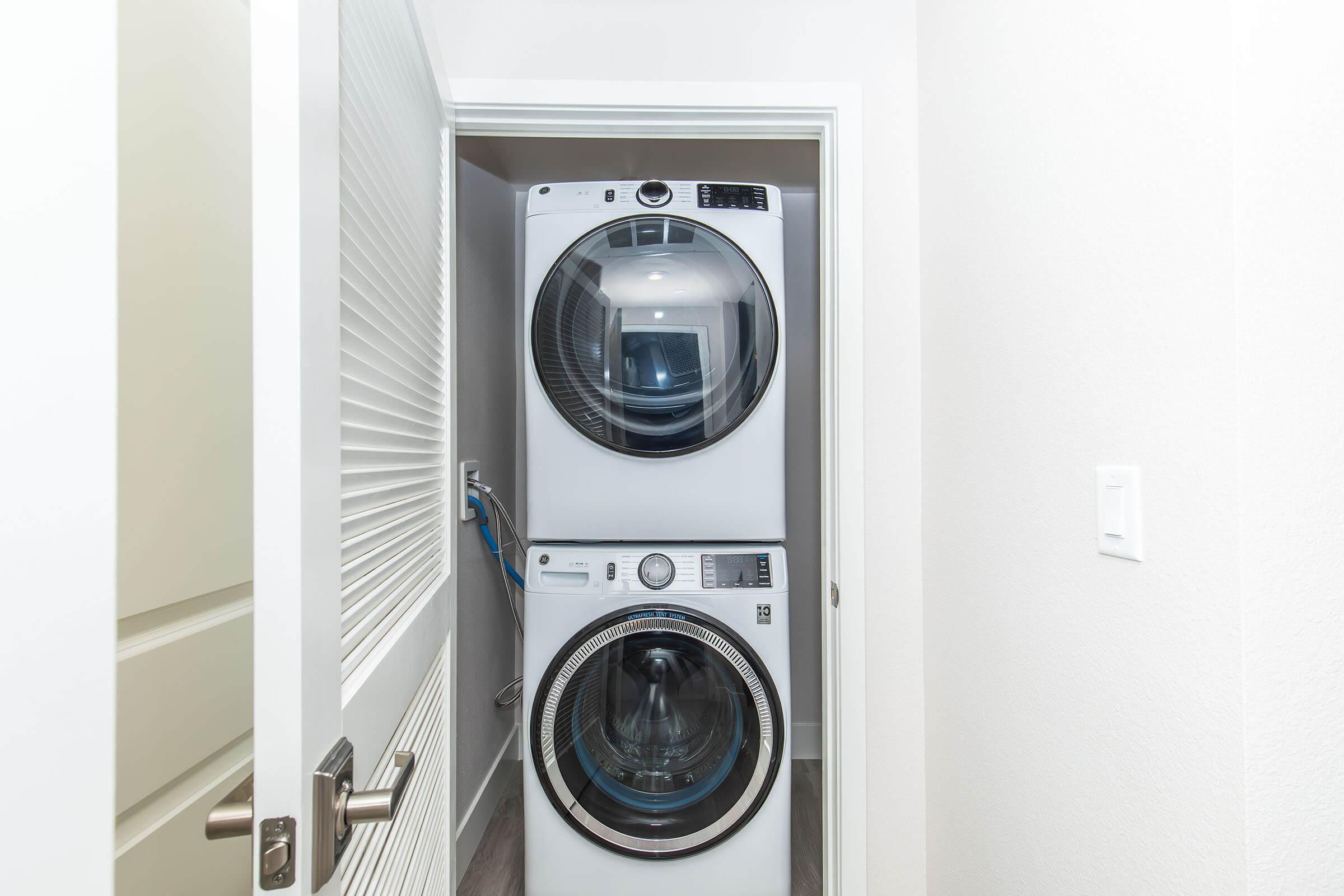 A modern laundry area featuring a stacked washer and dryer unit in a narrow closet space. The appliances are white, with glass doors, and the surrounding walls are light-colored. A partially open door reveals a clean and organized space.