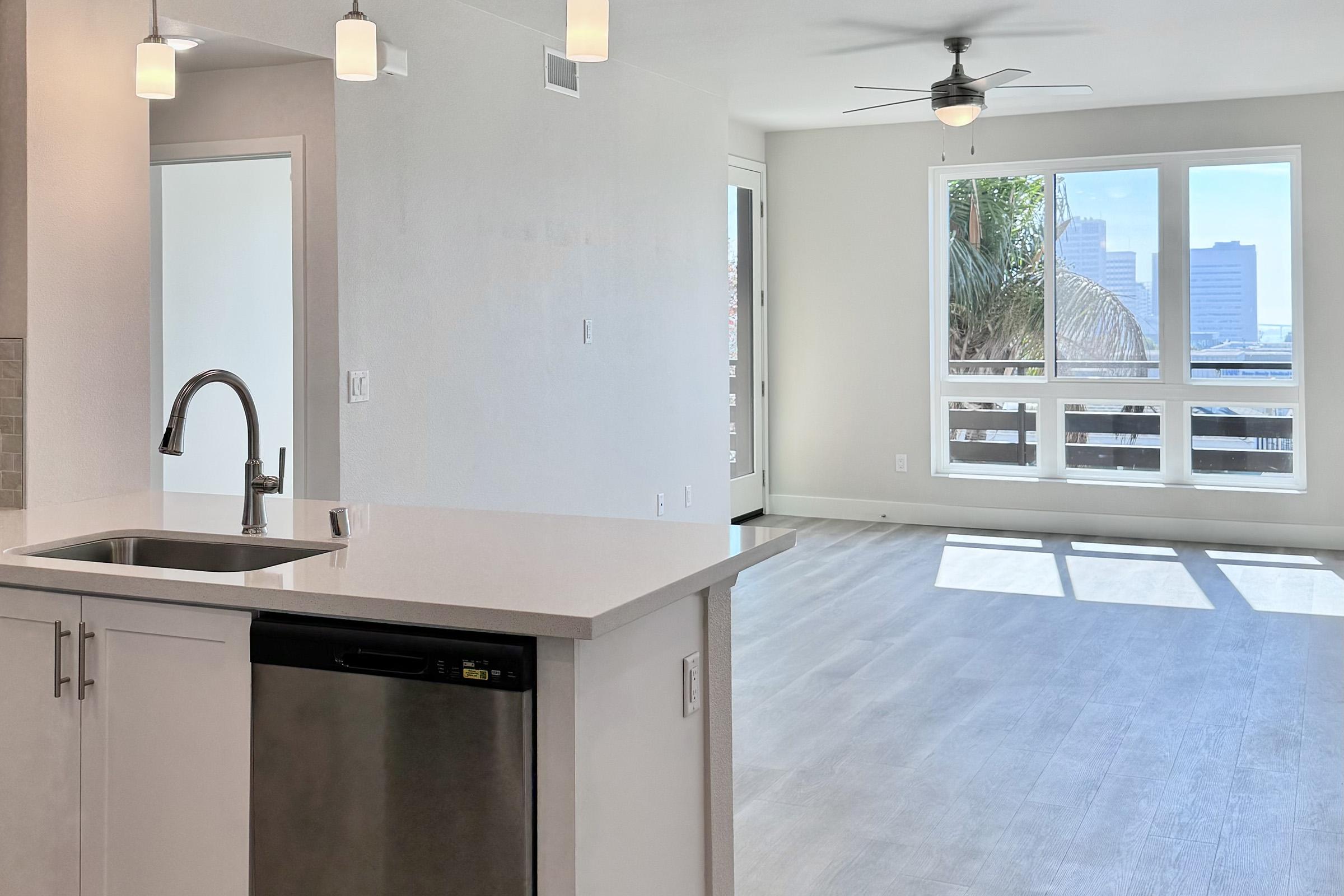 Bright and modern interior of an apartment featuring a kitchen area with a stainless steel sink and dishwasher. The living space has large windows allowing natural light, showing a view of city buildings and greenery outside. Light-colored flooring and walls create an open, airy atmosphere.