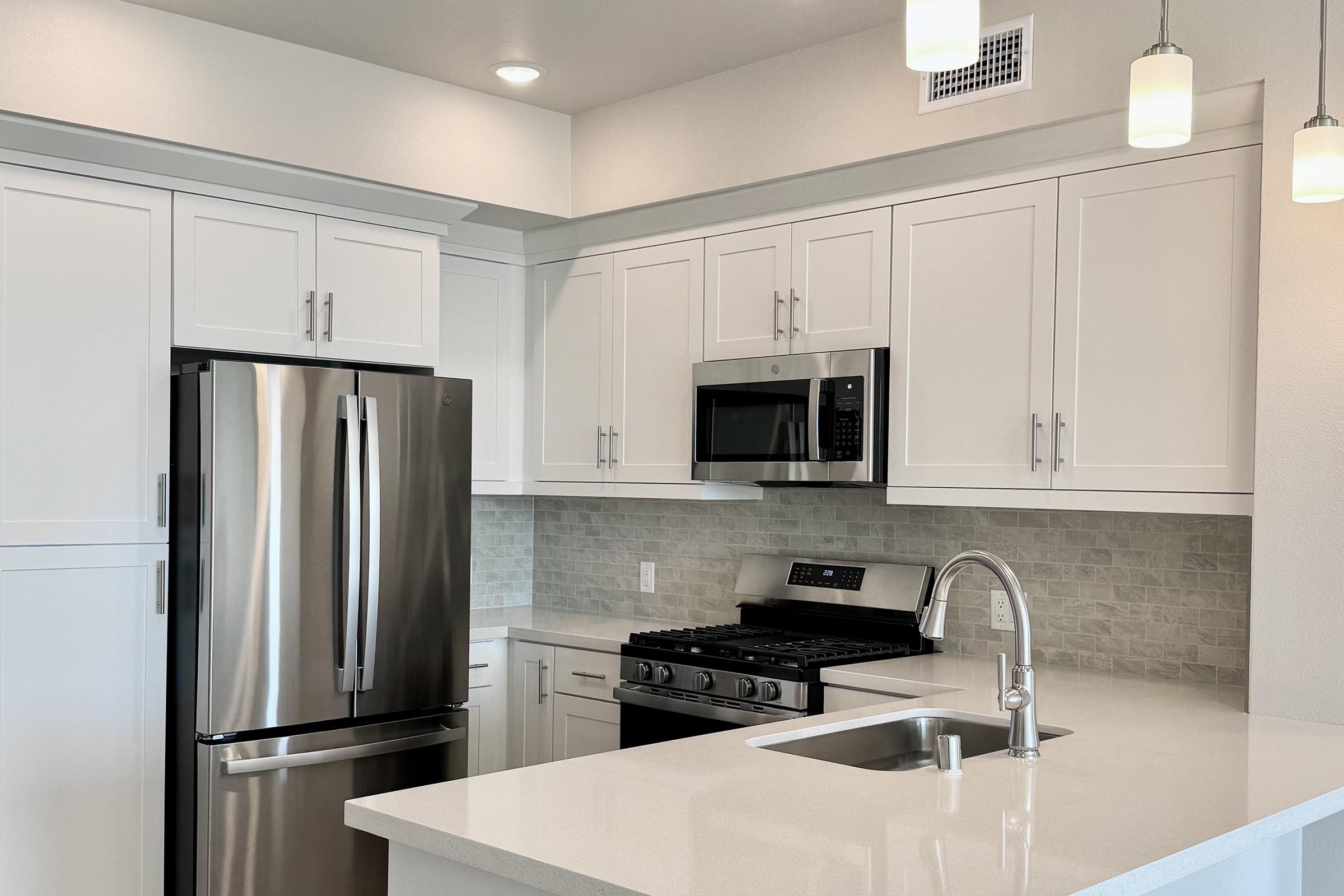 Modern kitchen featuring white cabinetry, stainless steel appliances including a refrigerator and microwave, a gas stove, and a sleek white countertop. The backsplash has light tiles, and pendant lights hang above the workspace, creating a bright and contemporary atmosphere.