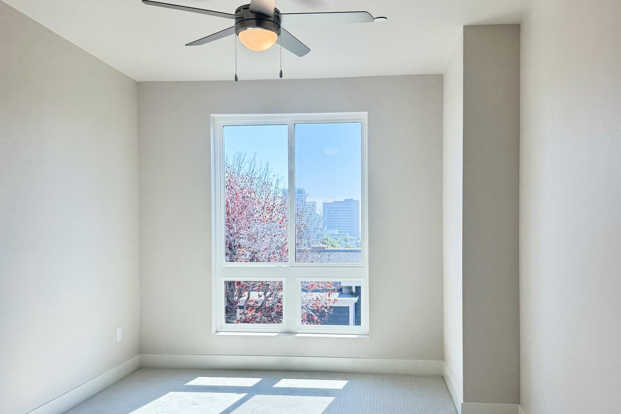 A bright, empty room with light-colored walls and a ceiling fan. Large windows reveal a view of trees and buildings outside, suggesting a sunny day. The floor is carpeted in neutral tones, and the space has a modern, minimalistic feel.