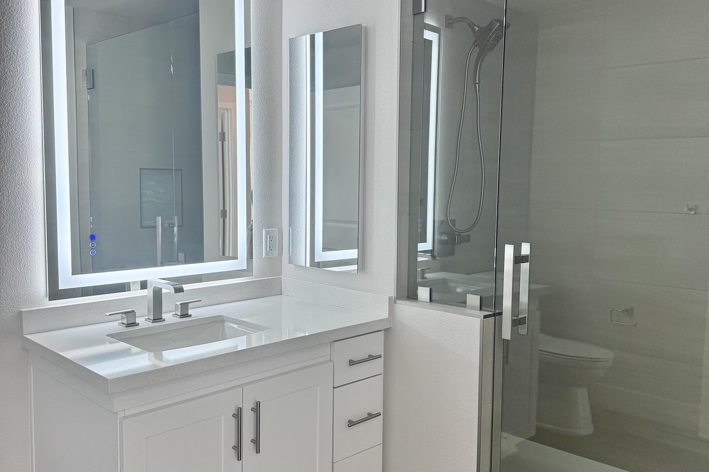 Modern bathroom featuring a sleek design with a vanity that has a white countertop and dual sinks. There are two illuminated mirrors above the sinks. A glass shower enclosure with a modern faucet is visible, alongside a white toilet. The overall color scheme is neutral, with bright lighting enhancing the space.