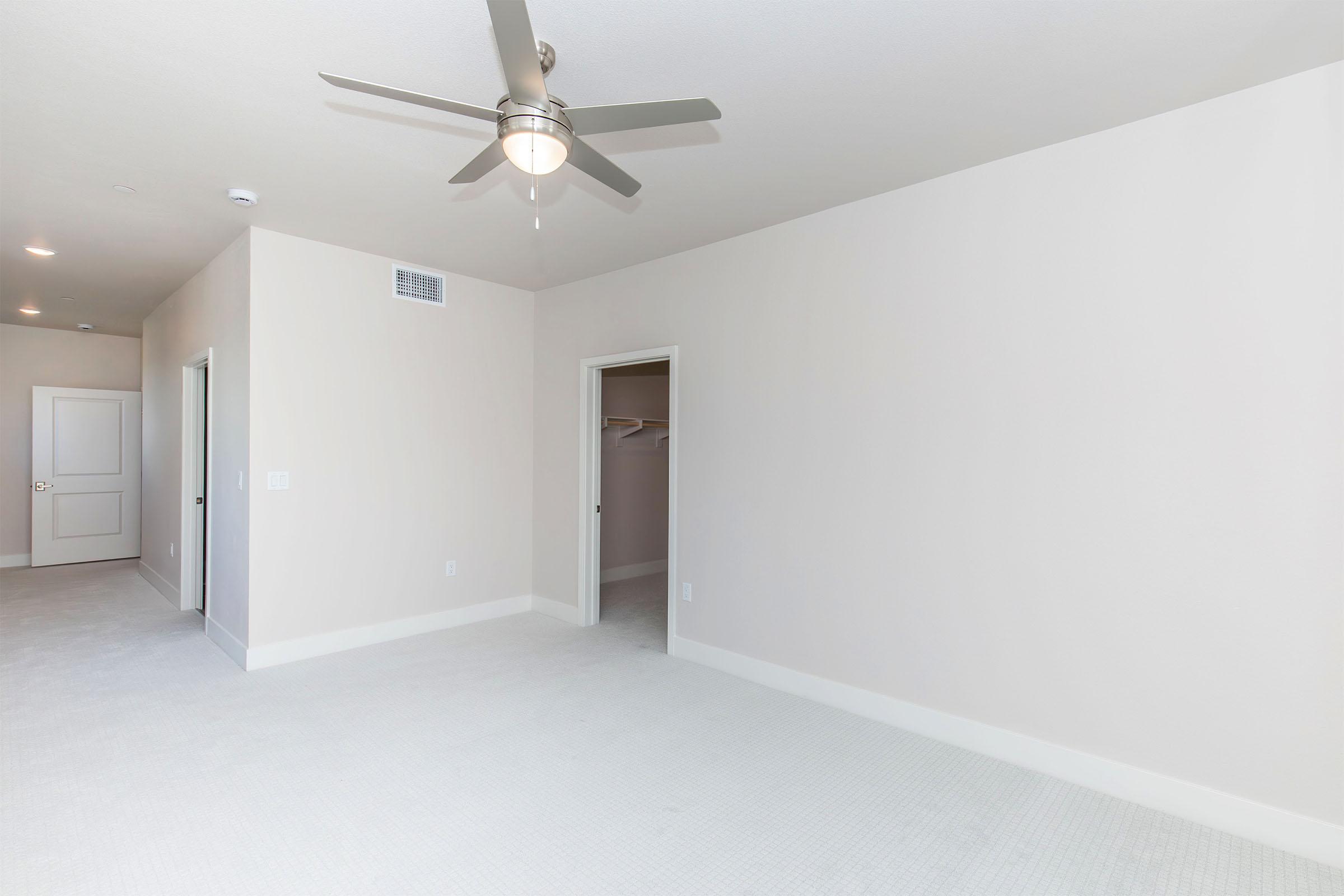 A spacious, empty room with light-colored walls and a ceiling fan. It features a doorway on the left leading to another room and an open closet area in the back. The floor is covered with a plush carpet, creating a clean and bright atmosphere. Ideal for various decorating possibilities.