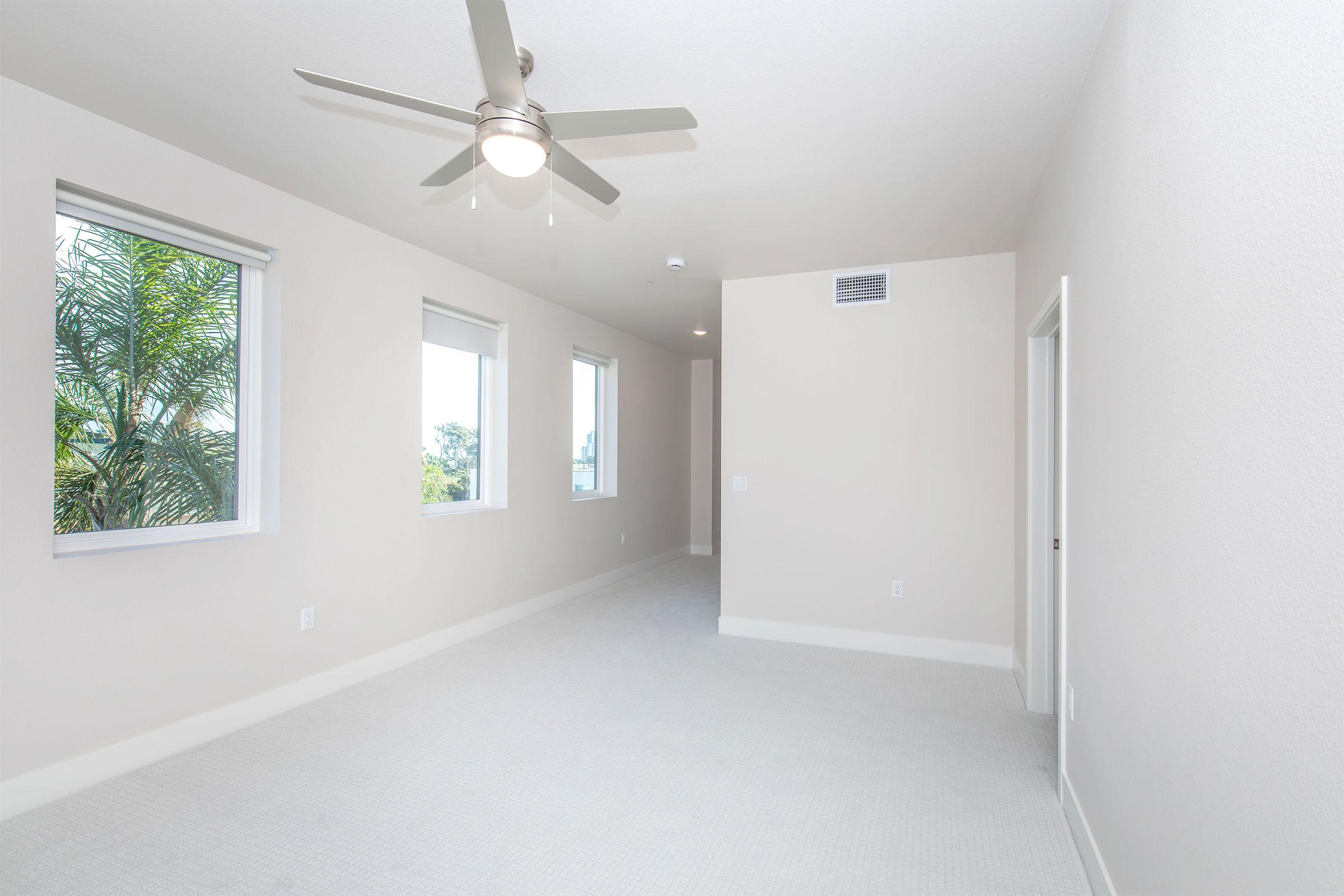 Spacious, well-lit hallway featuring light-colored walls and a carpeted floor. There are large windows allowing natural light and a ceiling fan for ventilation. The end of the hallway leads to a doorway, with a view of greenery outside visible through the windows.
