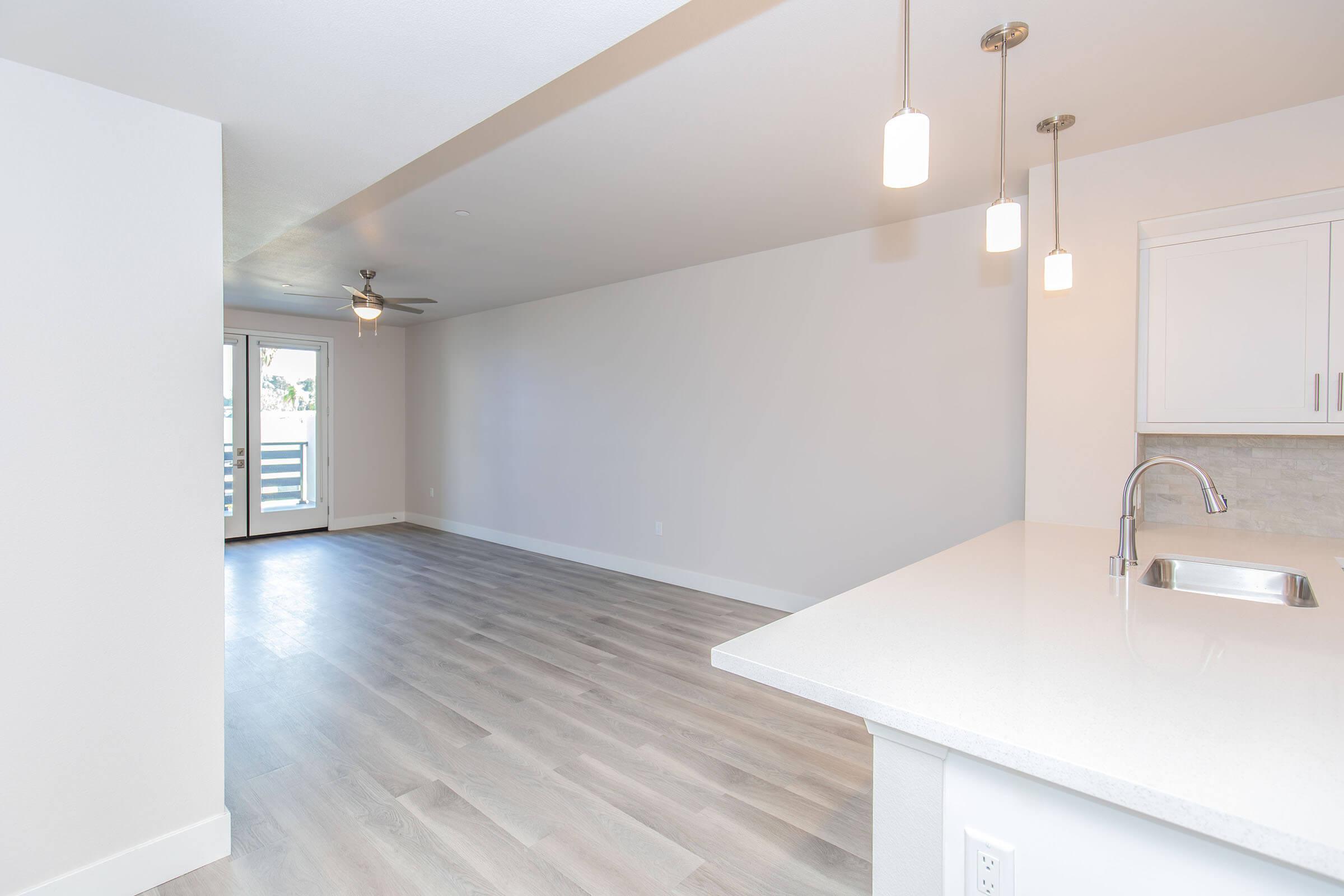 A bright and modern interior of an apartment featuring light-colored walls, hardwood-like flooring, and a kitchen area with pendant lights. The space includes a sliding glass door leading to a balcony, providing natural light and an open layout that connects the living and dining rooms.