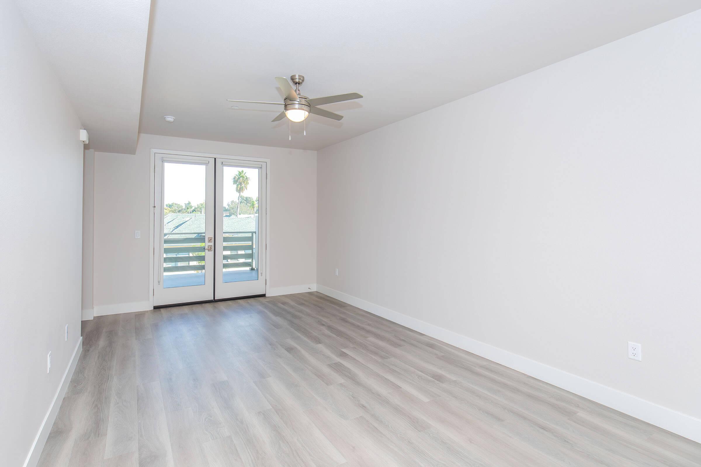An empty living room with light-colored walls and wooden flooring. It features a ceiling fan and has large sliding doors leading to a balcony. The space is well-lit with natural light coming in from the doors, creating a bright, airy atmosphere.