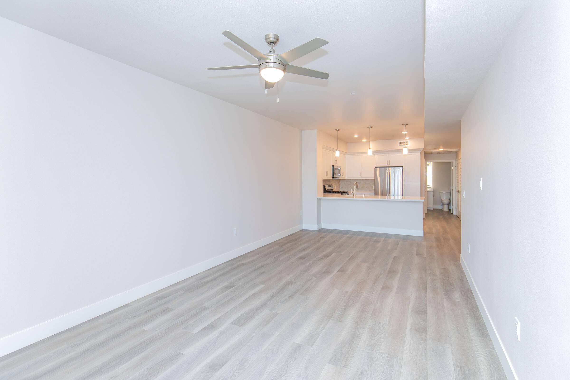 Contemporary living space featuring light gray walls and a ceiling fan. The room has laminate flooring and opens to a modern kitchen area with stainless steel appliances. Bright and airy atmosphere with ample natural light streaming in. Ideal for a cozy apartment setting.