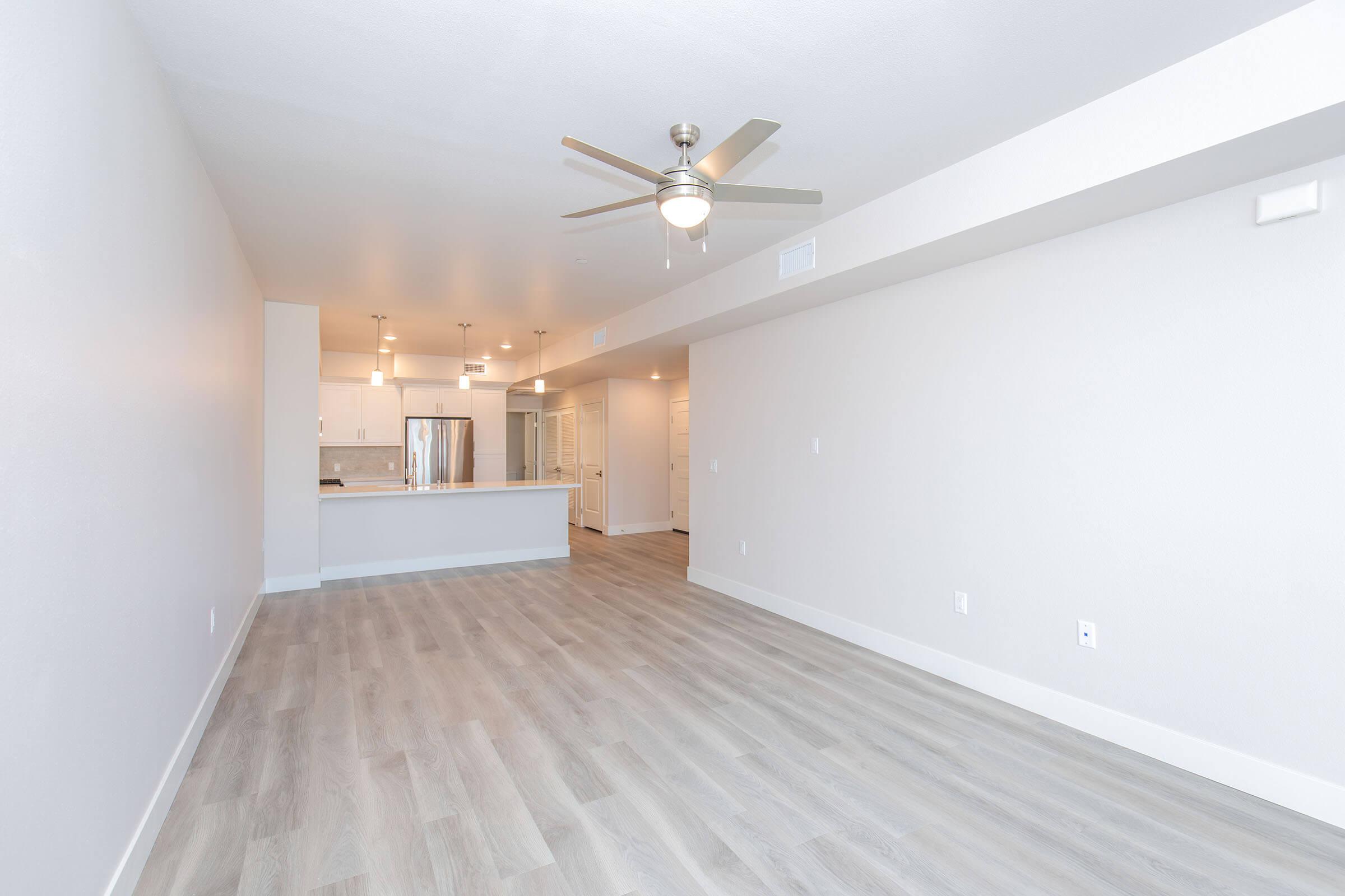 Bright and spacious modern apartment interior featuring open living area with light-colored hardwood flooring, white walls, and a ceiling fan. The kitchen is visible in the background, with stainless steel appliances and an island. Ample natural light fills the space, creating a welcoming atmosphere.