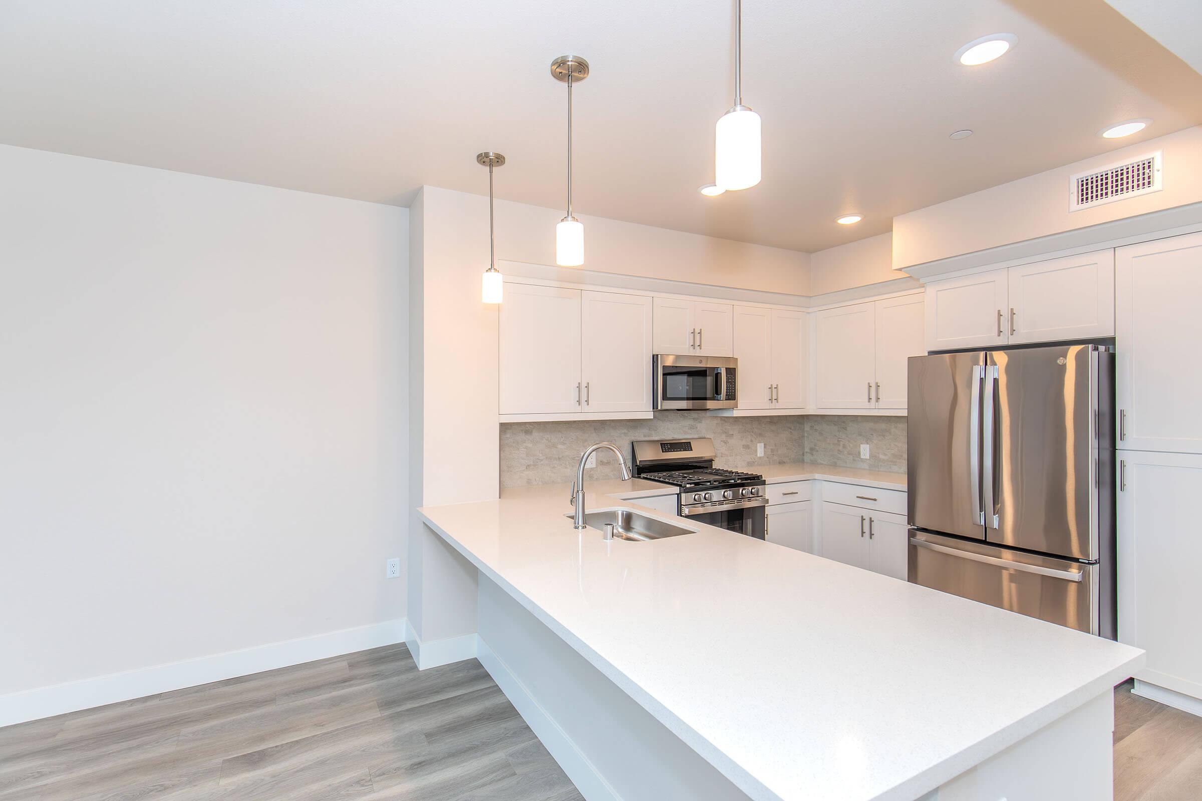 Modern kitchen featuring white cabinetry, a large island with a sink, stainless steel appliances including a refrigerator and oven, and stylish pendant lighting. The flooring is light wood, and the walls are painted in a neutral color, creating a bright and airy atmosphere.
