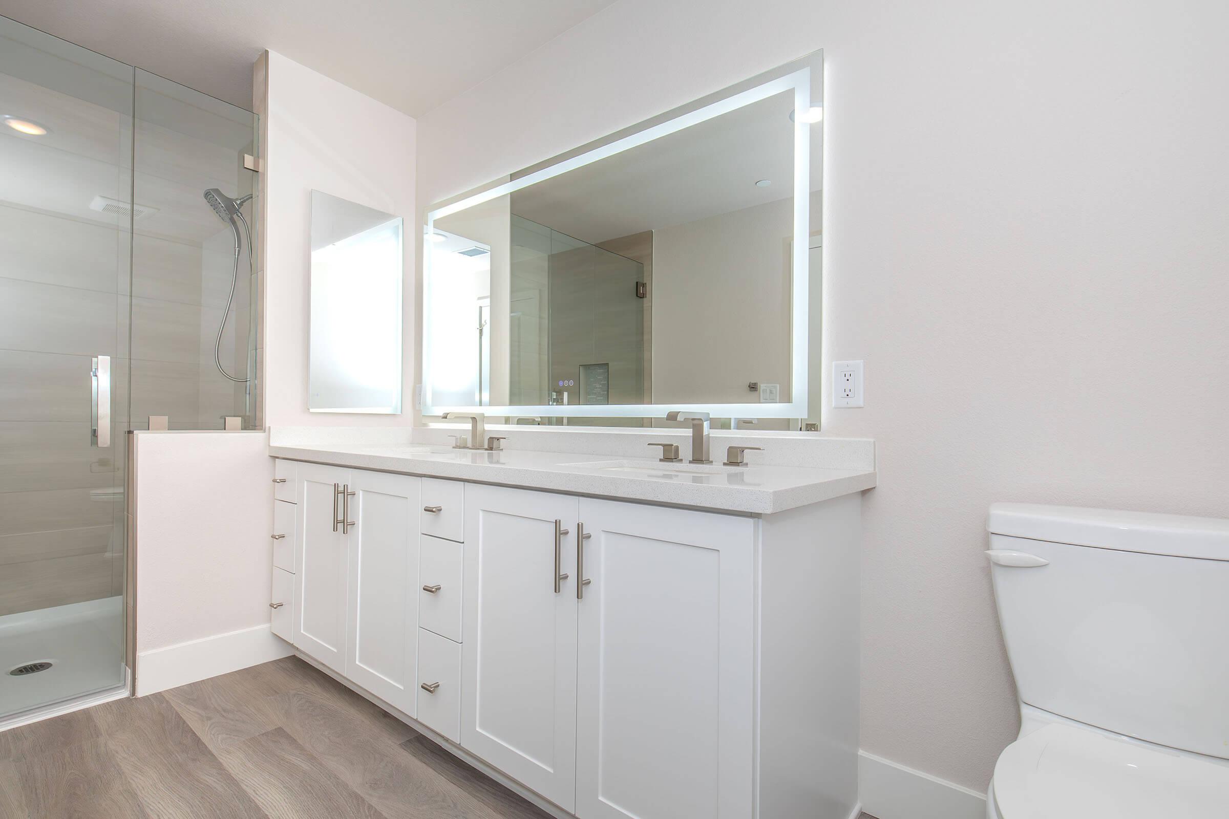 A modern bathroom featuring a double vanity with white cabinets and a countertop, two sinks, a large illuminated mirror, a glass shower enclosure, and a white toilet. The walls are a neutral color, and the flooring is light wood. Natural light enters through a window.