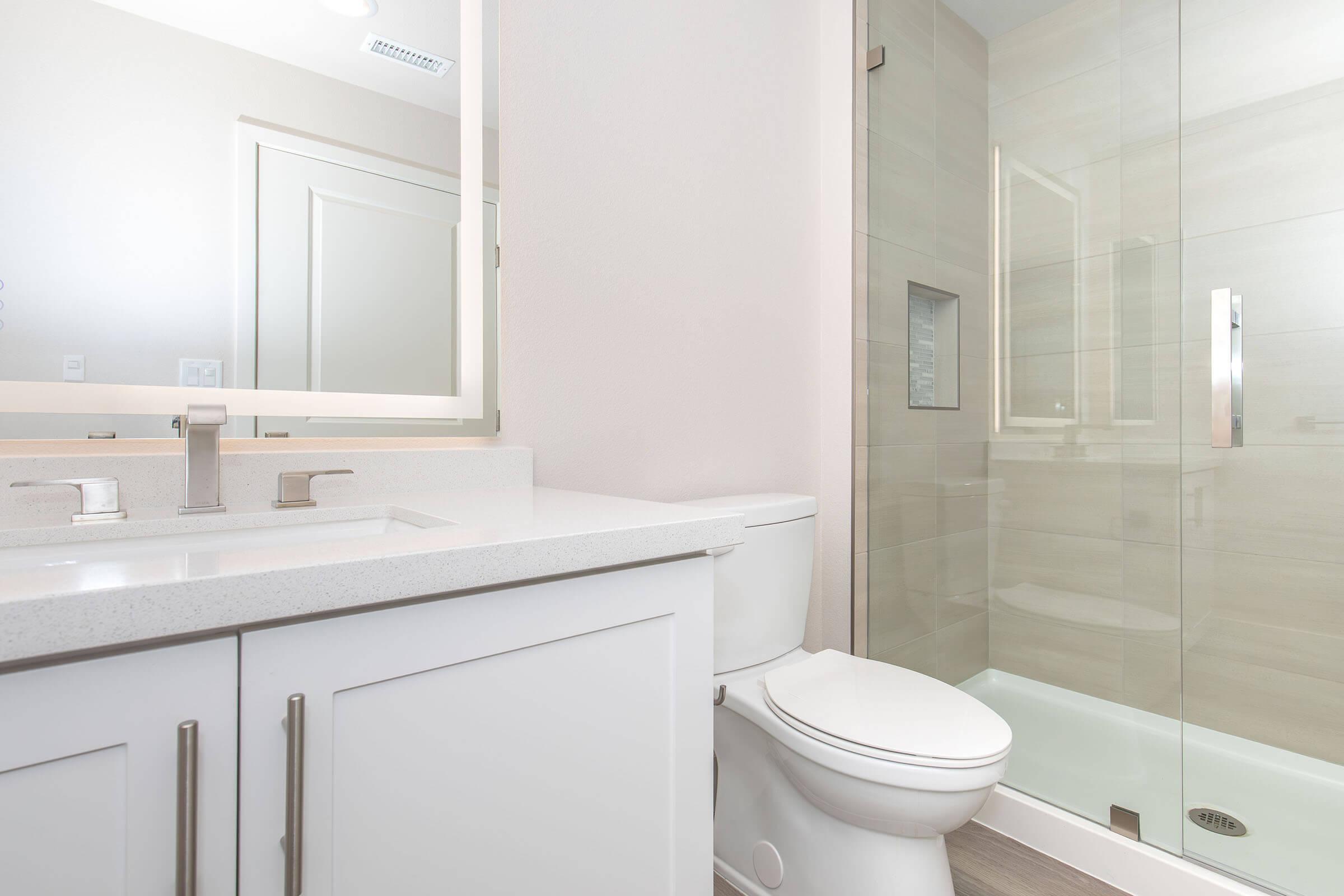 Modern bathroom featuring a white vanity with a sink, a toilet, and a glass shower enclosure. The walls are a light neutral color, and the space is well-lit with natural light reflecting off the surfaces. A minimalist design emphasizes cleanliness and contemporary aesthetics.