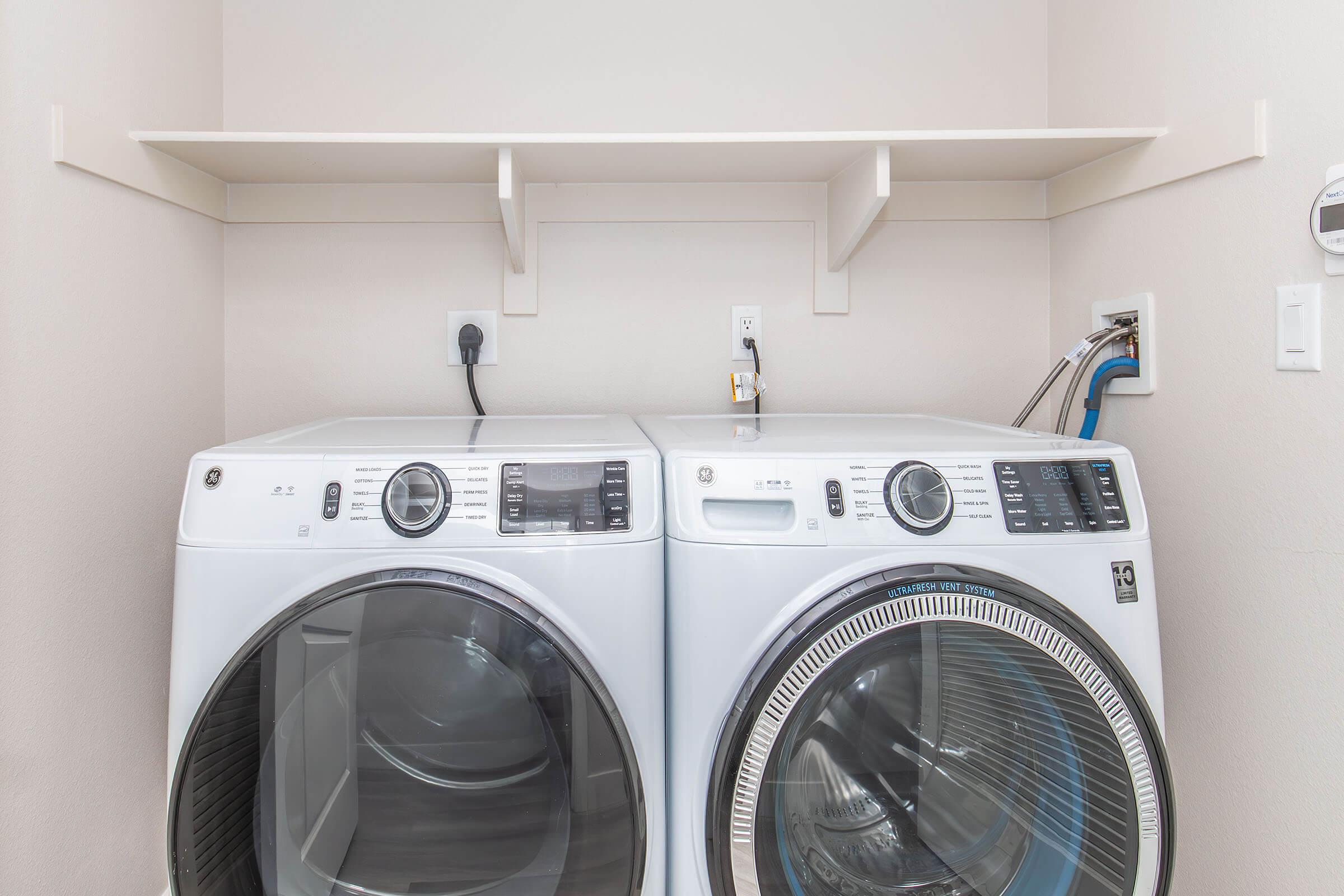 A pair of modern white washing machine and dryer set side by side in a laundry room, with a clean, neutral wall and a small shelf above them. The appliances have digital control panels and are plugged into wall outlets, with visible water and drainage connections.