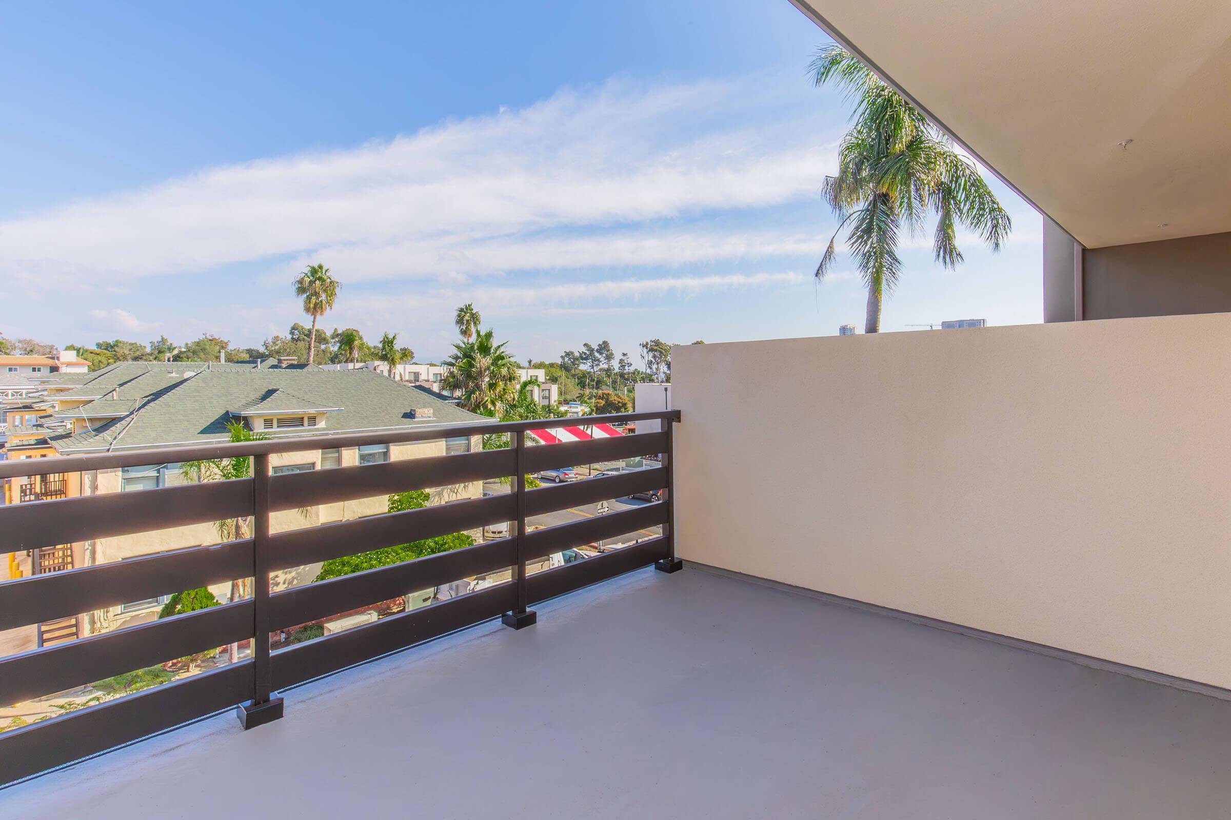 A sunny balcony with a modern railing, overlooking rooftops and palm trees. The clear blue sky features some scattered clouds, creating a bright, inviting outdoor space.