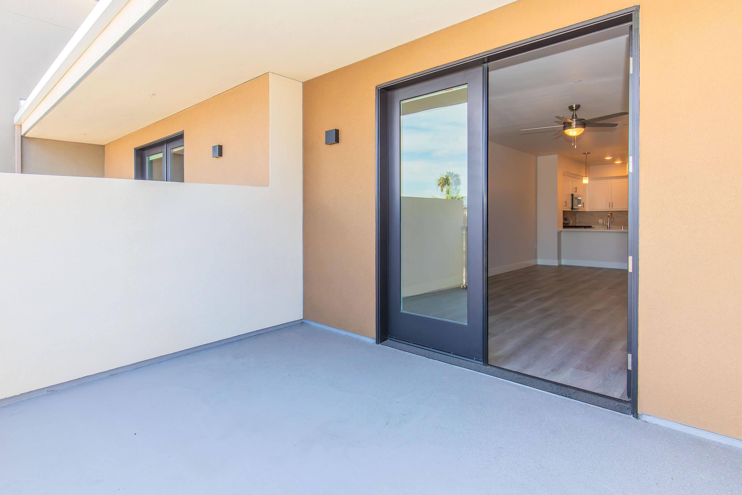 A modern balcony view of an apartment with sliding glass doors leading into an open living space. The interior features light-colored walls, stylish lighting, and a visible kitchen area. The balcony floor is smooth and the walls are a warm beige, giving a contemporary feel.