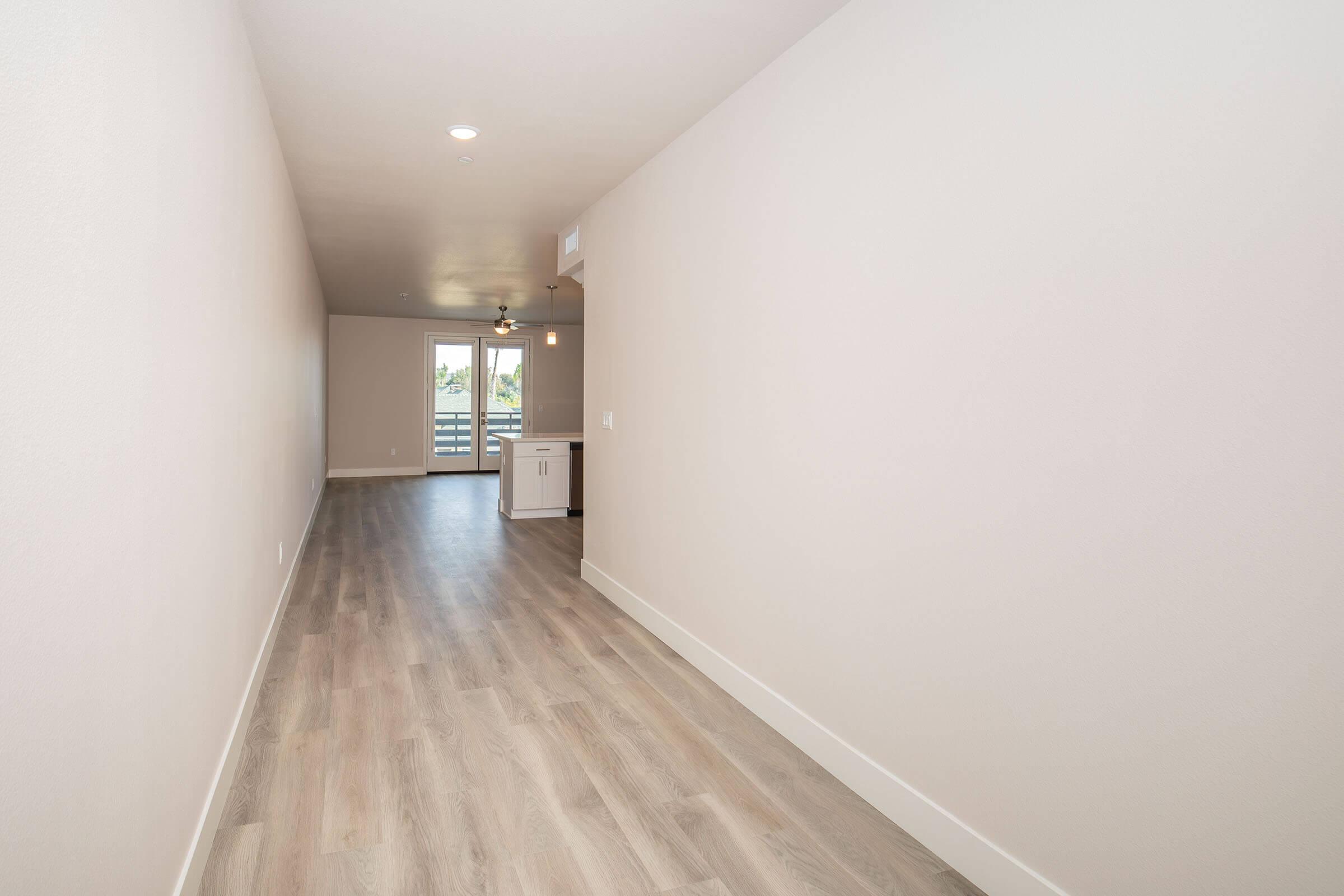 A spacious hallway with light-colored walls and light wood flooring, leading to a doorway with sliding glass doors that open to a balcony. The area is well-lit with a ceiling fan visible, and a small kitchen area is partially seen at the end of the hallway.