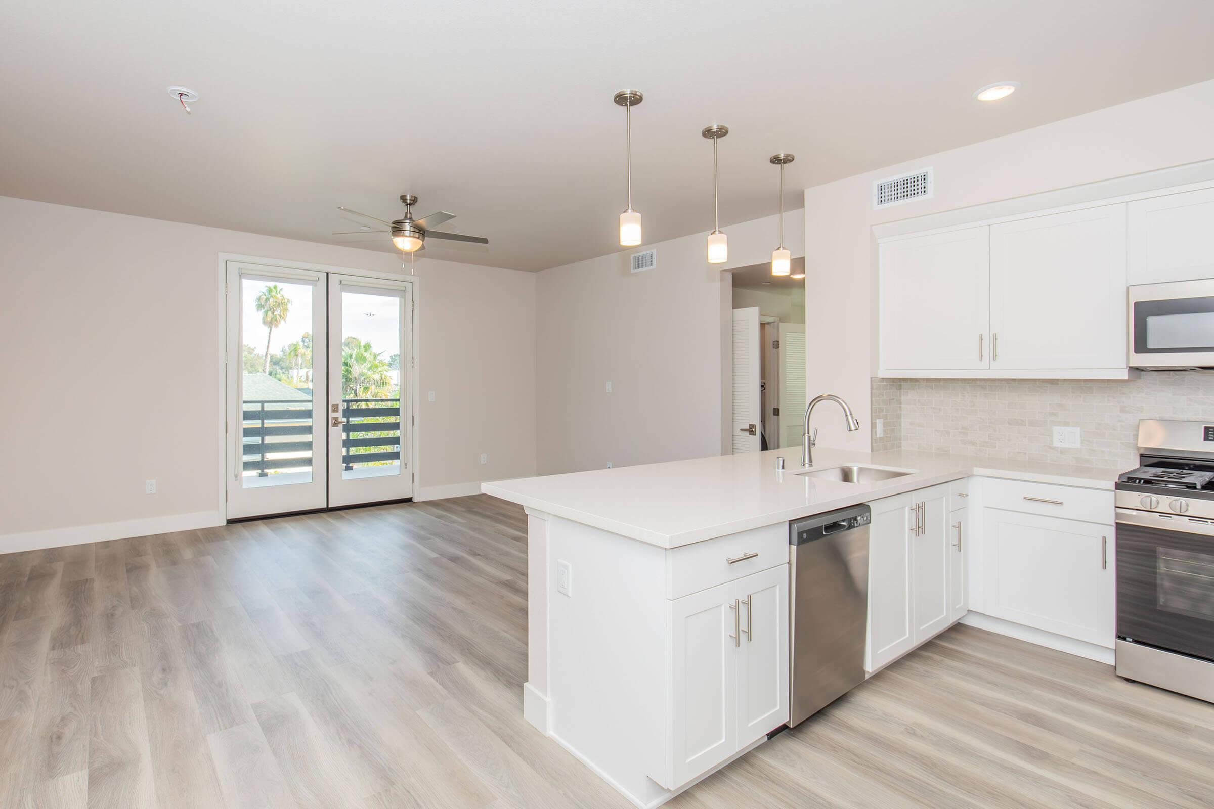 Modern kitchen and living area in an apartment with light wood flooring. The kitchen features white cabinets, stainless steel appliances, a large island with a sink, and pendant lighting. There are sliding glass doors leading to a balcony, with natural light brightening the space.