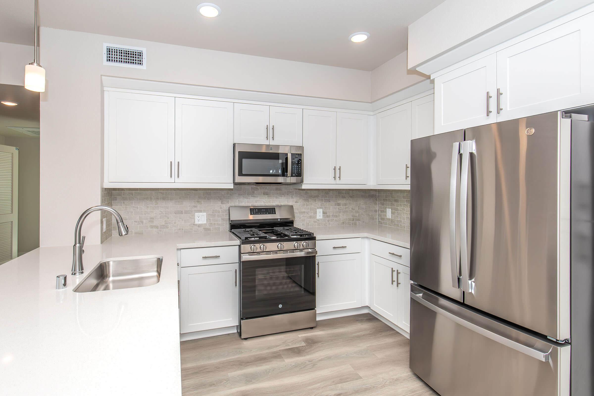 Modern kitchen featuring white cabinetry and stainless steel appliances. Includes a gas stove, microwave, and a large refrigerator. The countertop is light-colored with a subtle texture, and there is a sleek sink and faucet. Soft lighting and a neutral color scheme create a bright and inviting atmosphere.