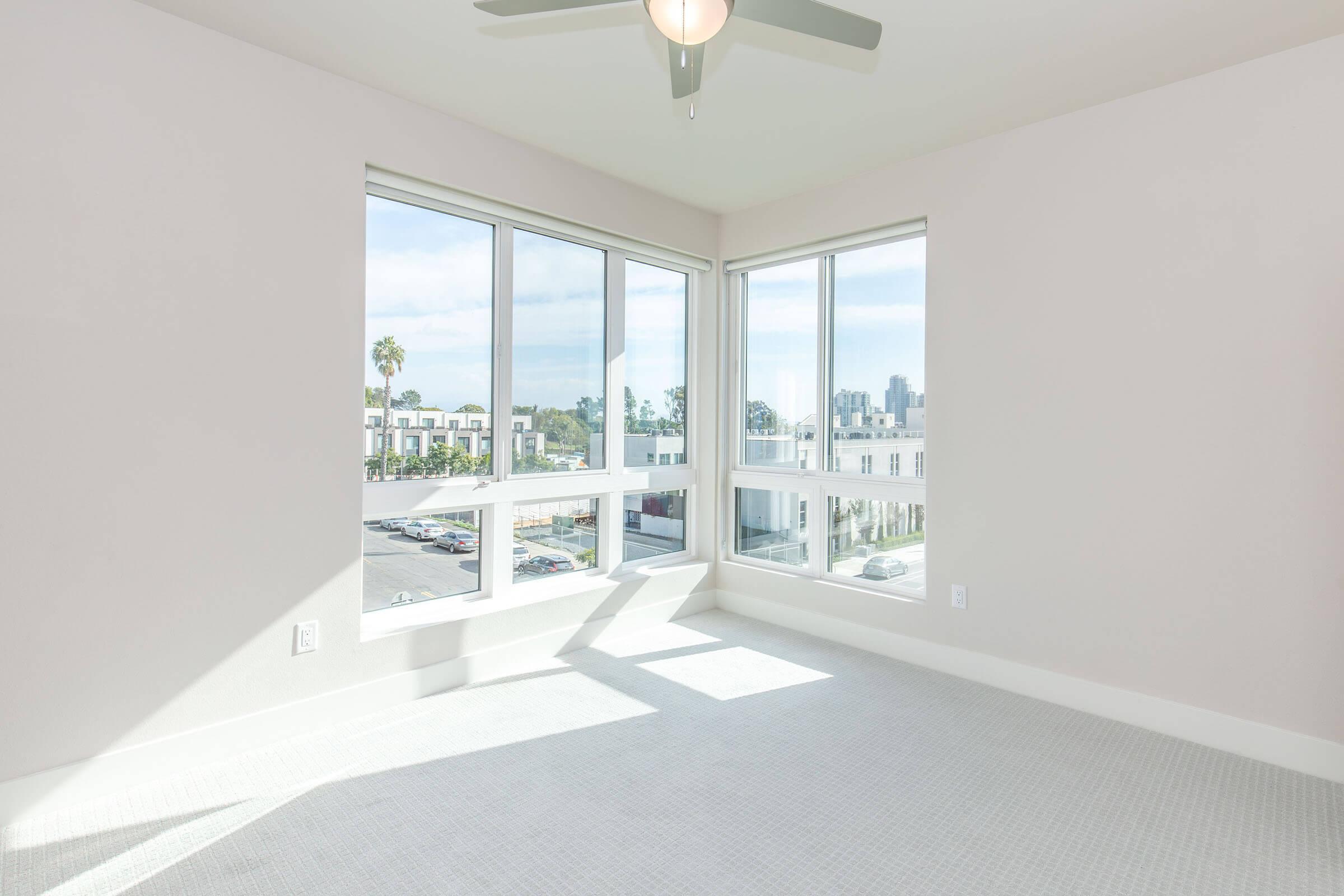 Bright, airy room with large windows allowing natural light, featuring a ceiling fan. The walls are painted in a soft color, and the flooring is light carpet. Outside the windows, there is a view of palm trees and a cityscape in the distance, creating a spacious and inviting atmosphere.