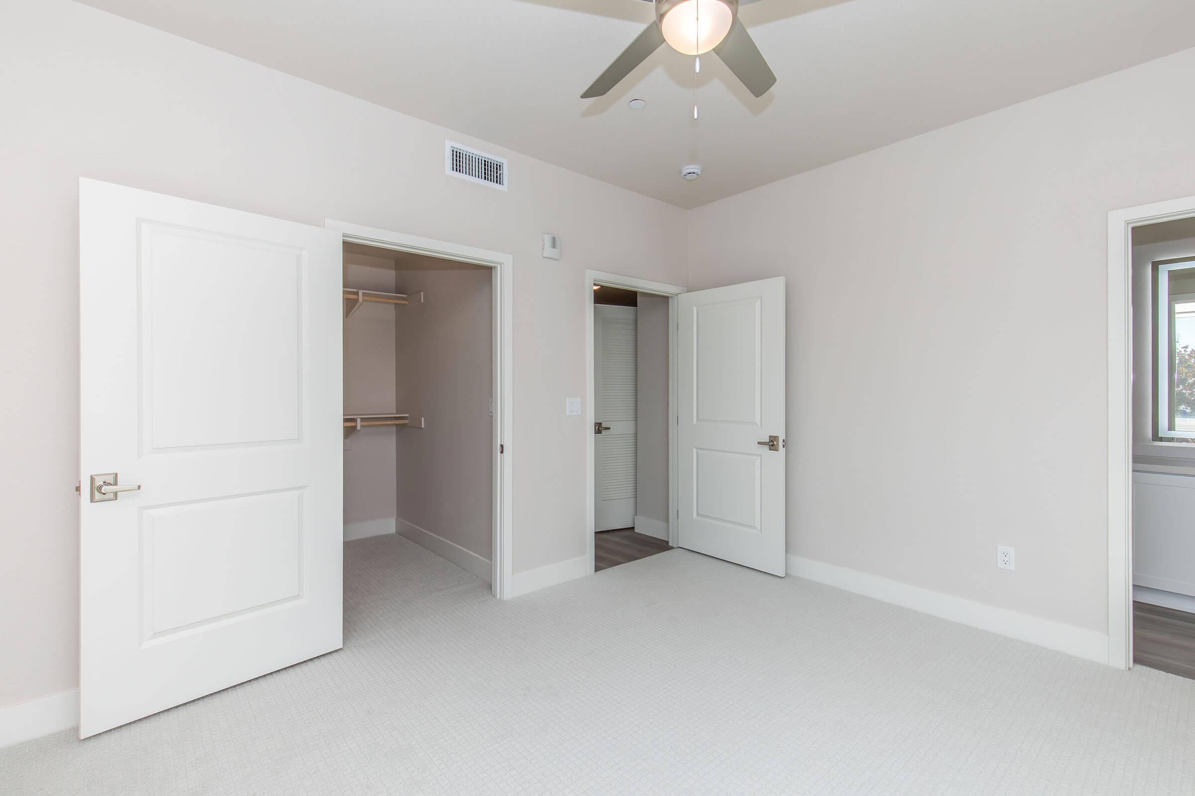 A bright, empty bedroom featuring light-colored walls and a ceiling fan. There are two open doors, one leading to a closet space, and the other to an adjoining room. The floor is covered with soft carpeting, and large windows allow natural light to fill the space.