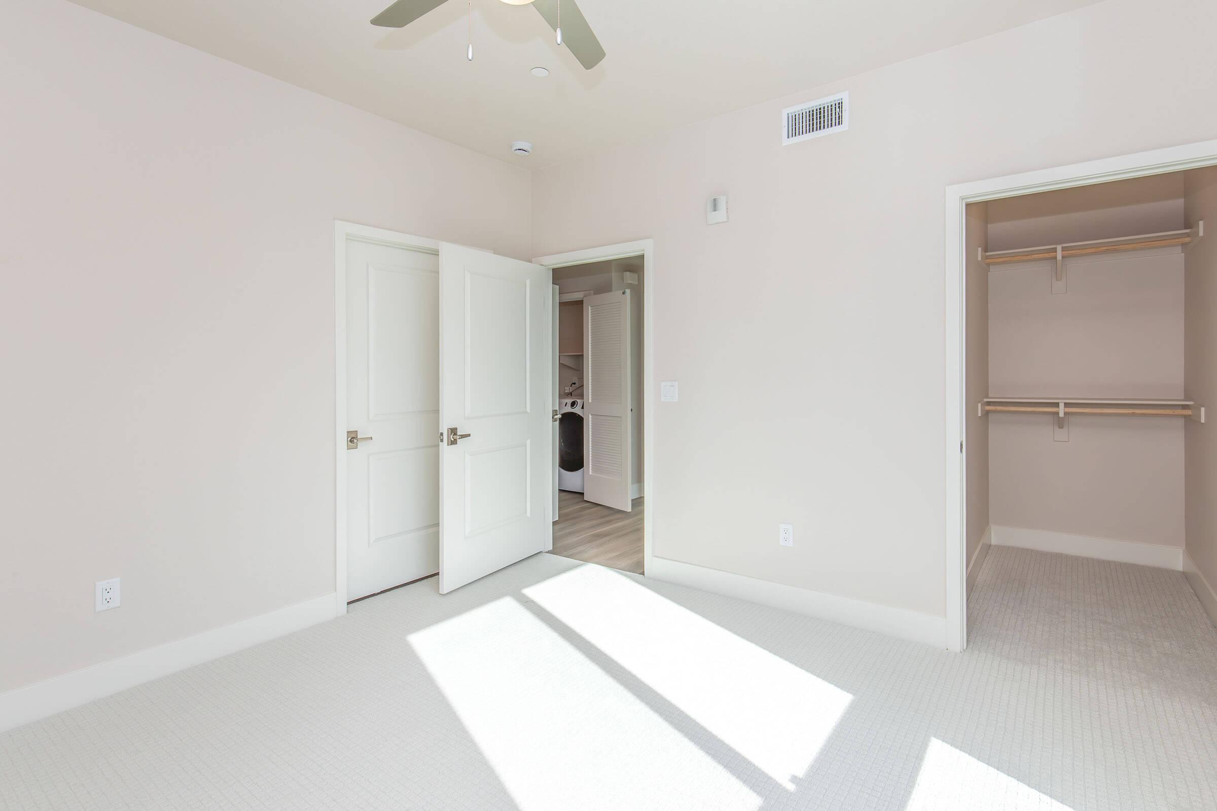 A spacious, empty room with light-colored walls and carpet. There are two open doors: one leading to a closet with wooden shelves and another possibly leading to a laundry area. Sunlight is streaming in, creating bright patches on the floor. A ceiling fan is mounted above.