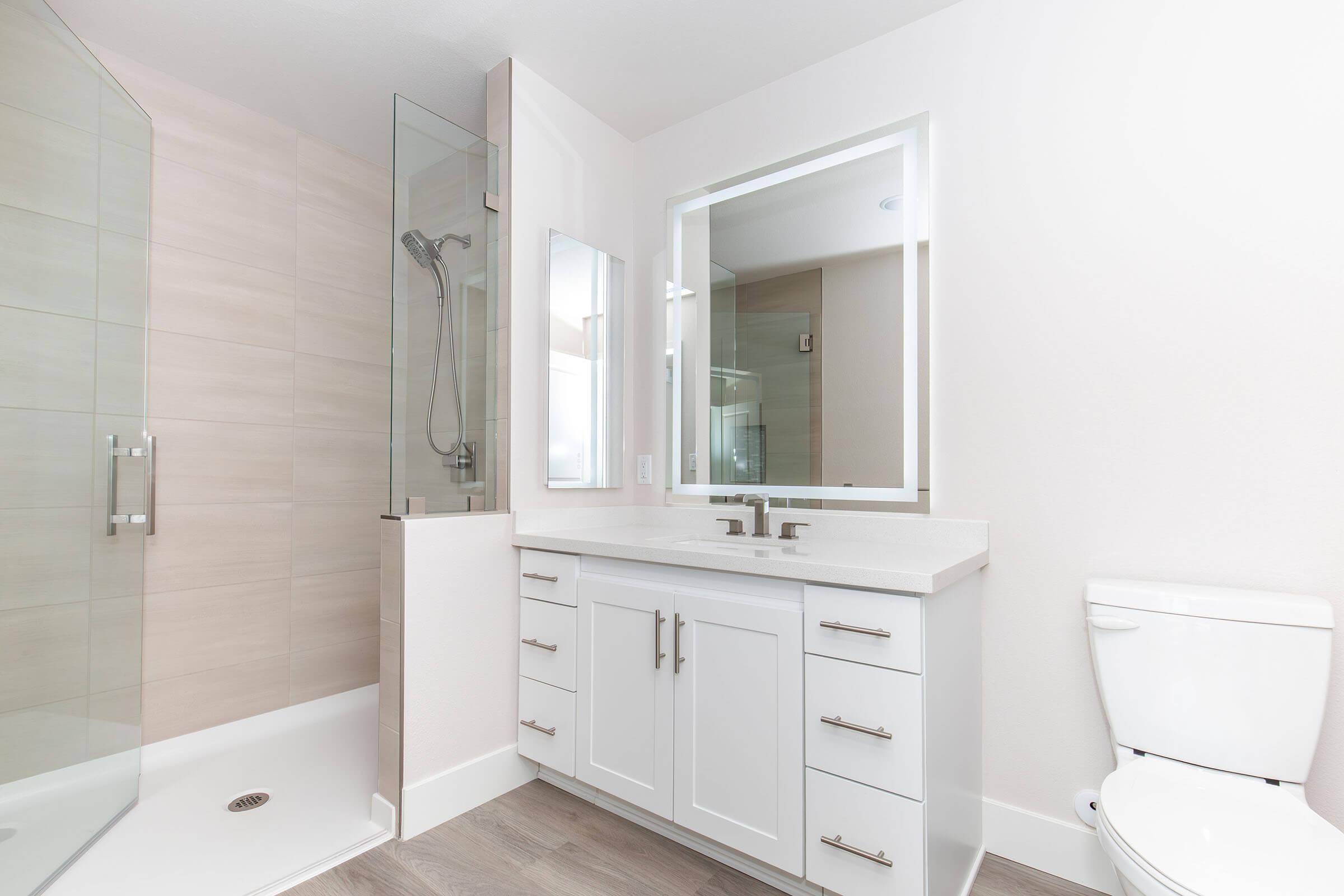 Modern bathroom featuring a glass shower enclosure, a large mirror above a double-sink vanity with white cabinets, and a white toilet. The walls are light-colored, and the flooring is a wood-like material, creating a clean and minimalist aesthetic.