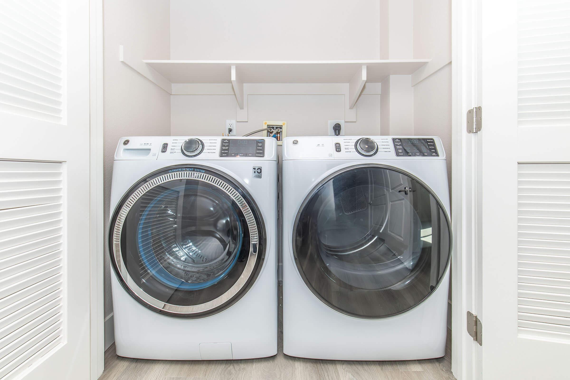 A modern laundry room featuring a pair of front-loading washing machines. The machines are white with transparent doors, neatly placed side by side in a compact space. Above them, there are two shelves for storage, and the walls are light-colored, creating a clean and organized look.