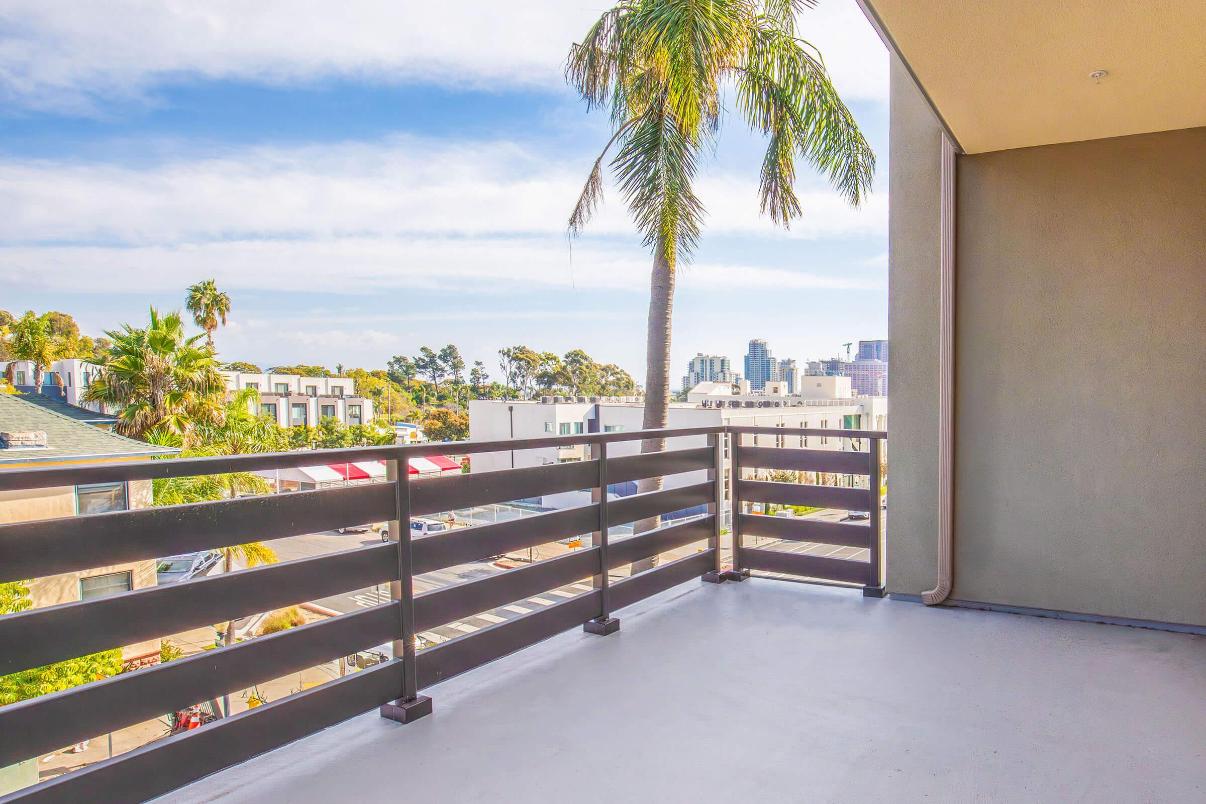A balcony view featuring a palm tree in the foreground, with city buildings visible in the background against a partly cloudy sky. The balcony railing is wooden, and the floor is smooth and clean. The scene suggests a bright, sunny day.