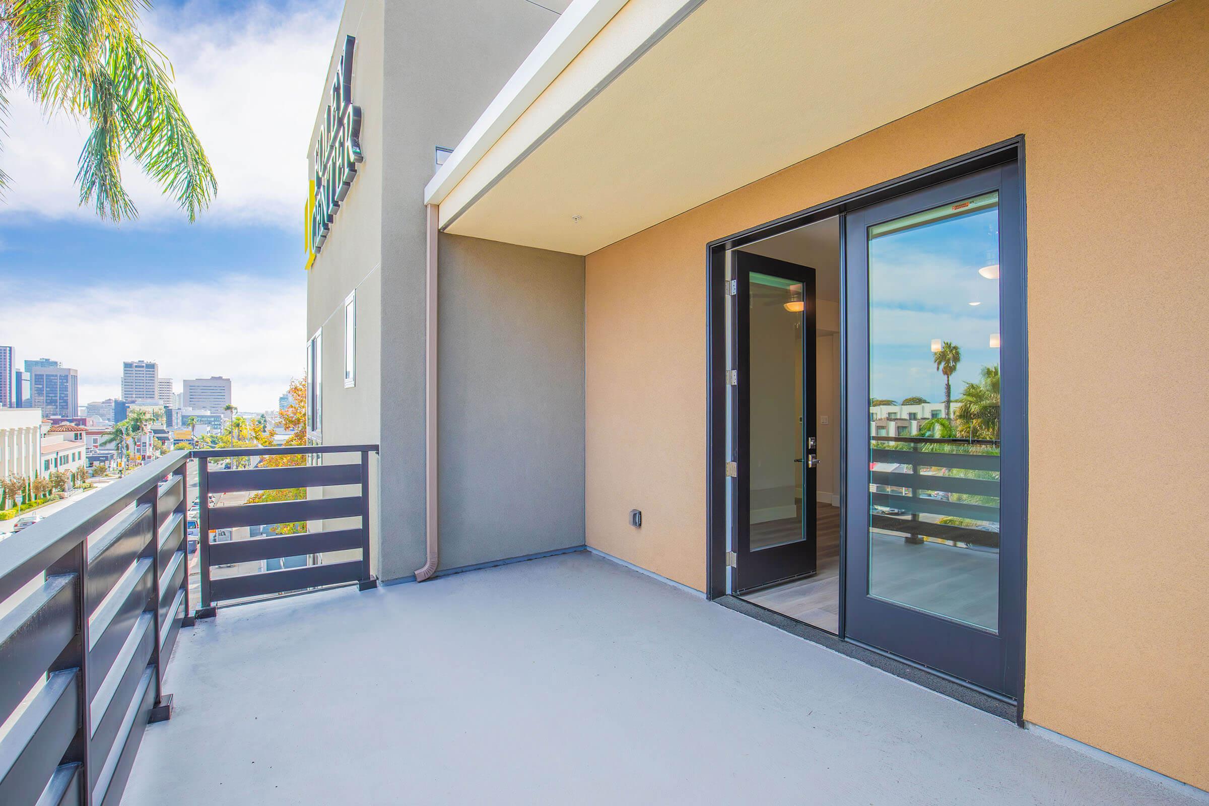 A modern balcony with a concrete floor and black railing, featuring a door leading to an interior space. The view includes palm trees and city buildings under a bright sky. The exterior walls are painted in light and dark tones, giving a contemporary look.