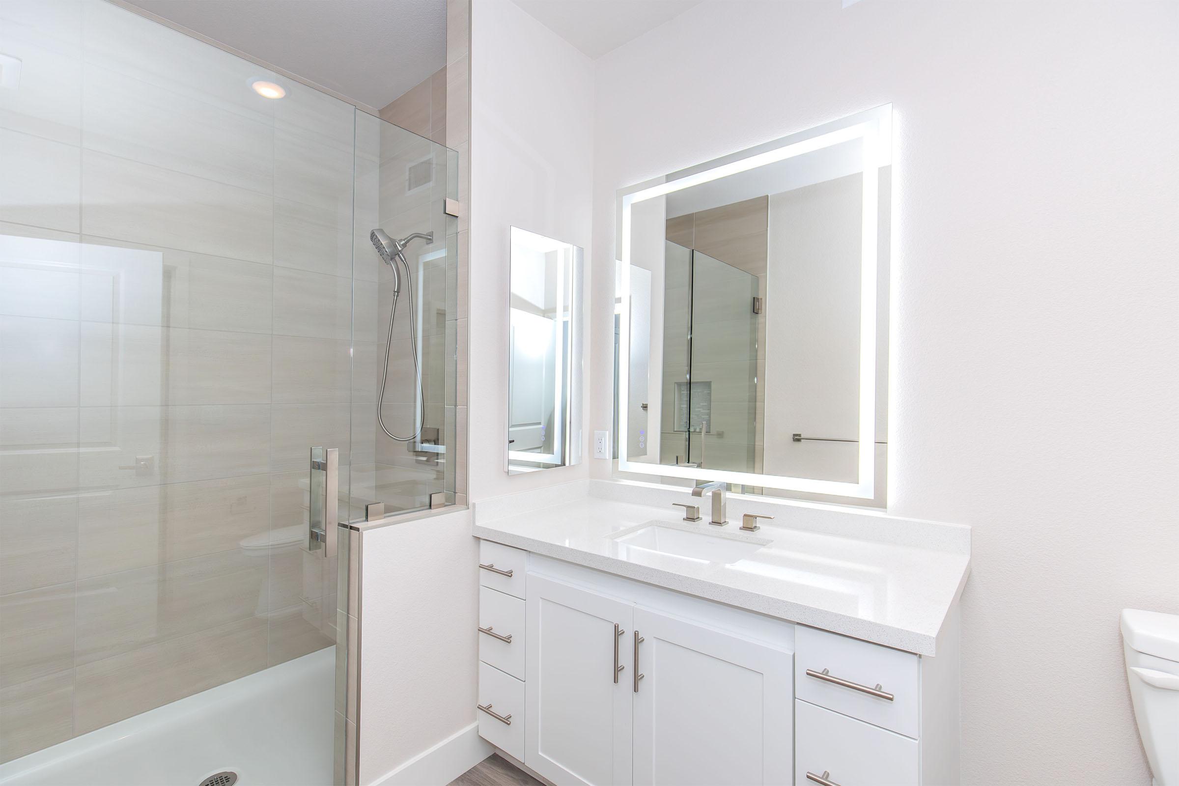 A modern bathroom featuring a glass shower enclosure, a sleek white vanity with two sinks, and a large mirror with integrated lighting. The walls are light-colored, and the flooring is gray, creating a clean and contemporary look. Natural light enters through a nearby window.