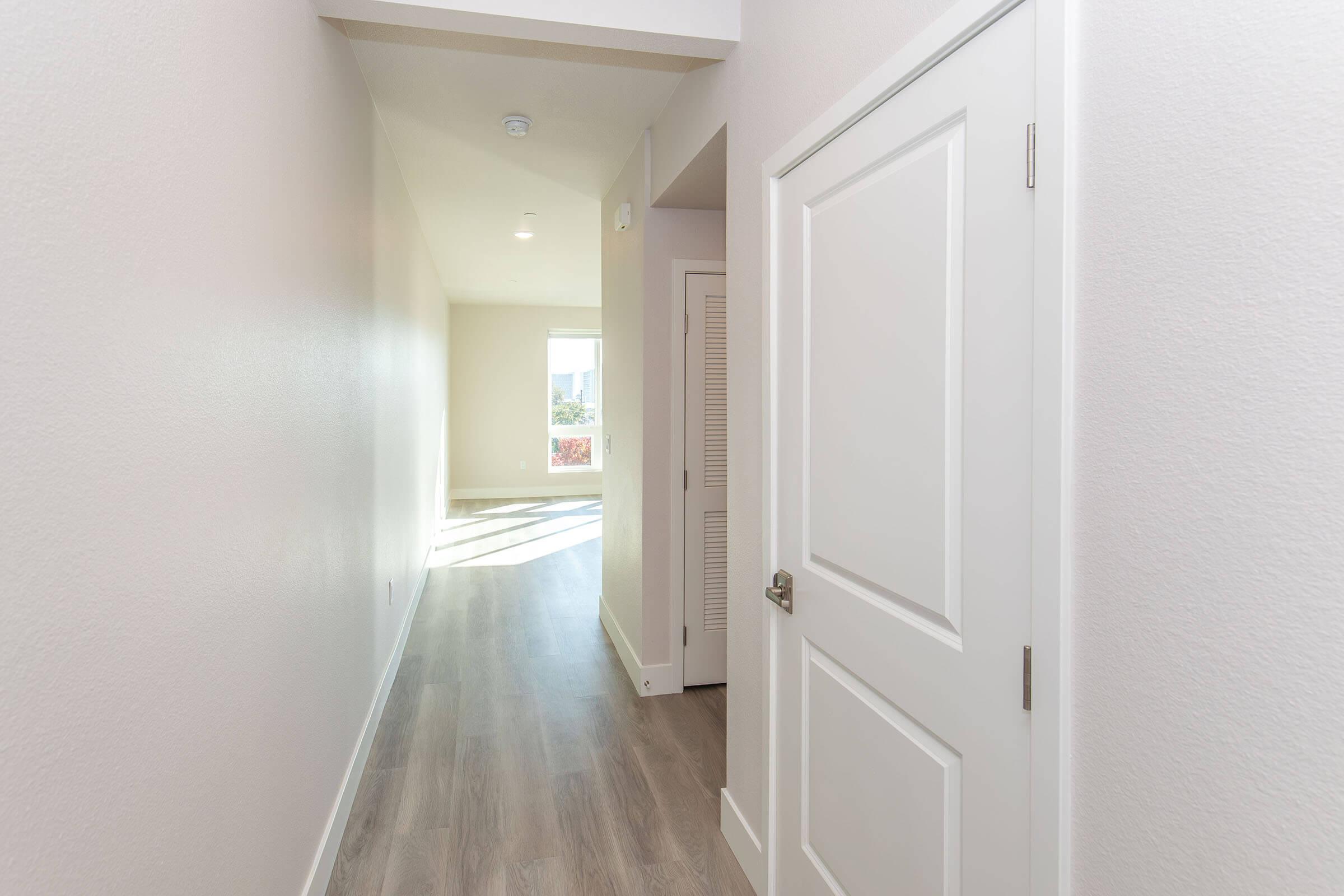 A well-lit hallway in a modern interior, featuring light-colored walls, a white door, and light wooden flooring. Natural light streams in from a window at the end of the hallway, creating an inviting atmosphere. The space appears clean and freshly painted.