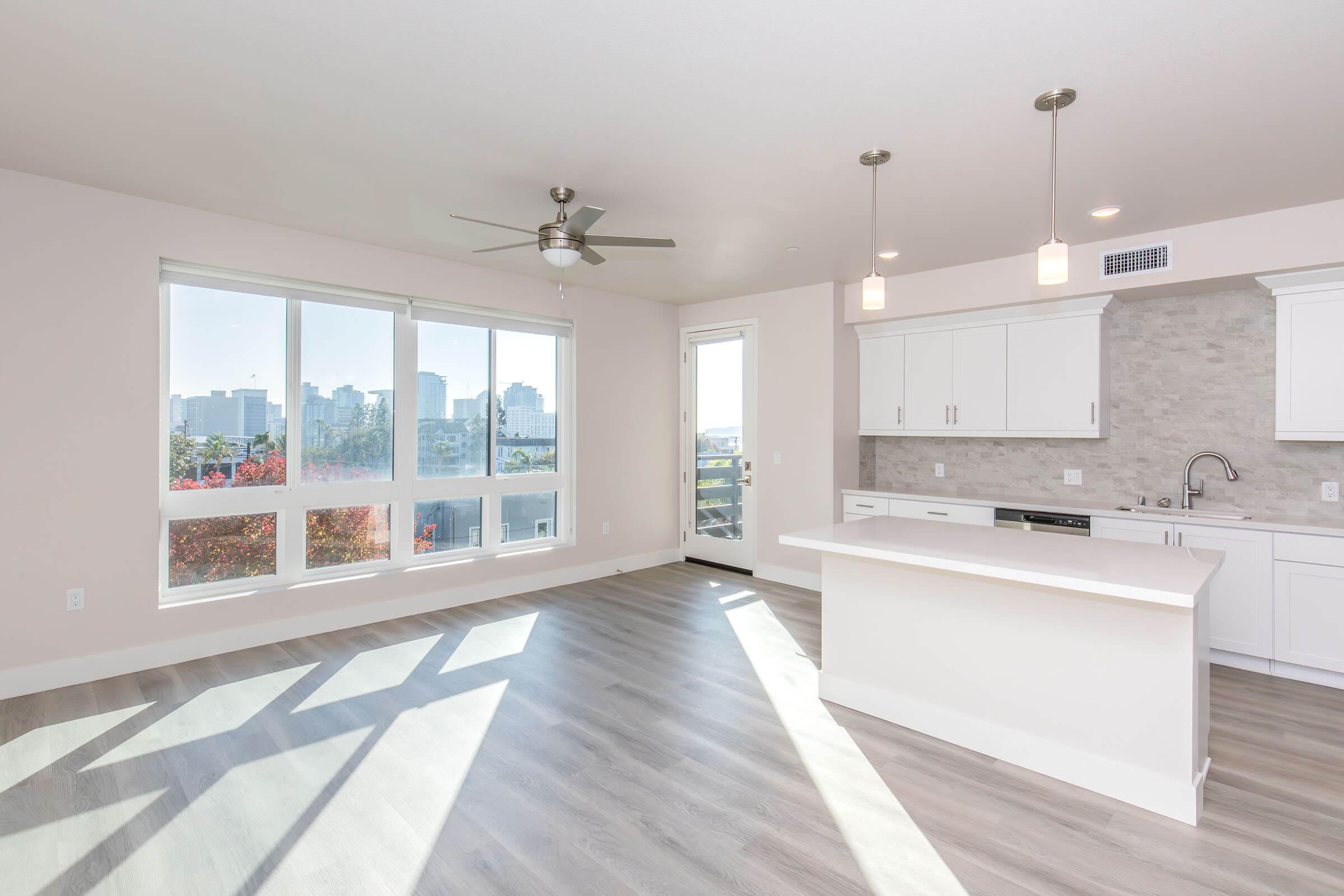 A modern, empty living space with large windows allowing natural light, offering a view of city buildings. The room features light-colored walls, wood-style flooring, and a white kitchen area with minimalistic cabinets and a countertop. A ceiling fan is installed, enhancing the open, airy feel.