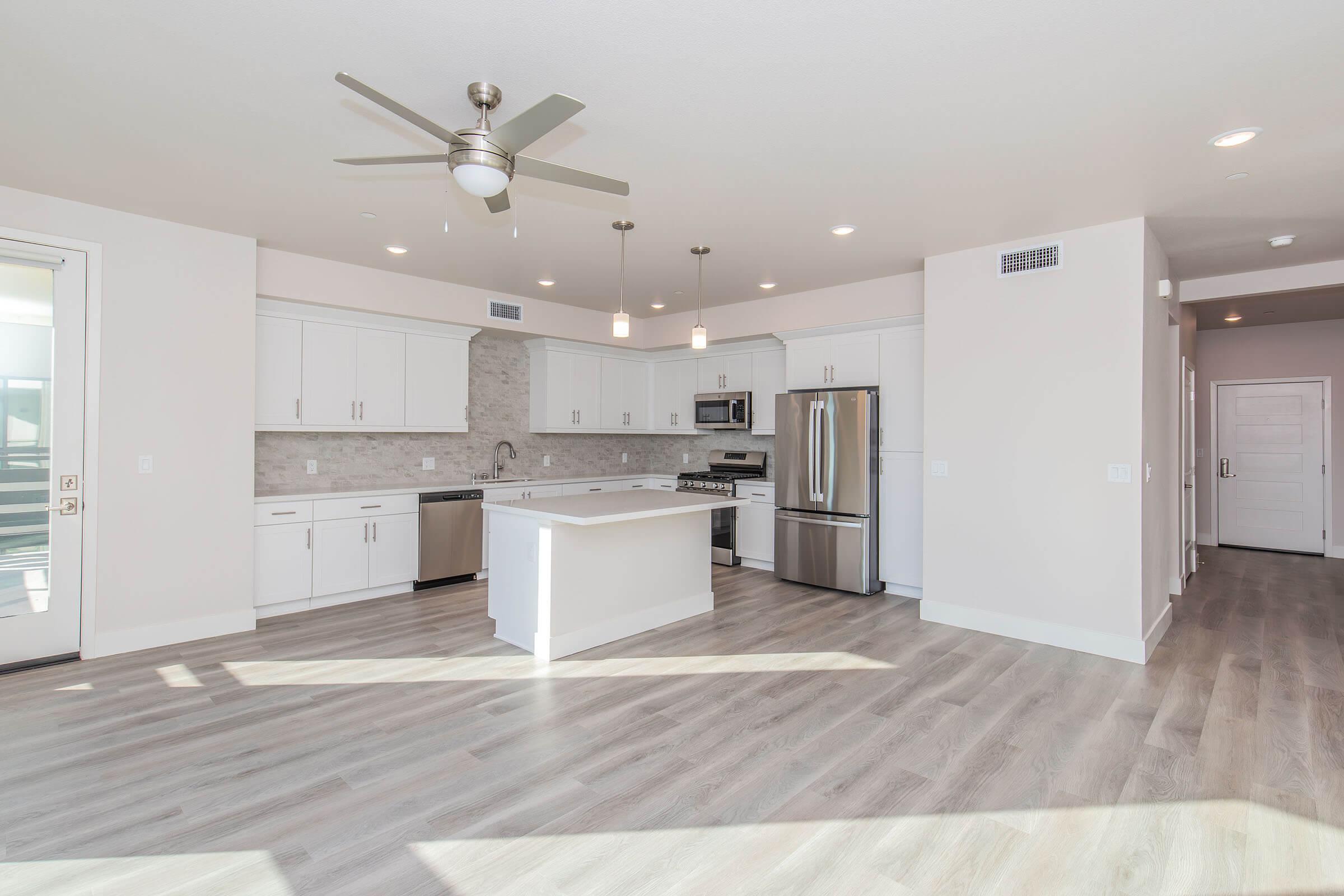 Modern kitchen and living area featuring light wood flooring, white cabinets, stainless steel appliances, and a large center island. Bright, spacious layout with overhead lighting and a ceiling fan. Wall of windows and a door leading to an outdoor space, creating an inviting atmosphere.