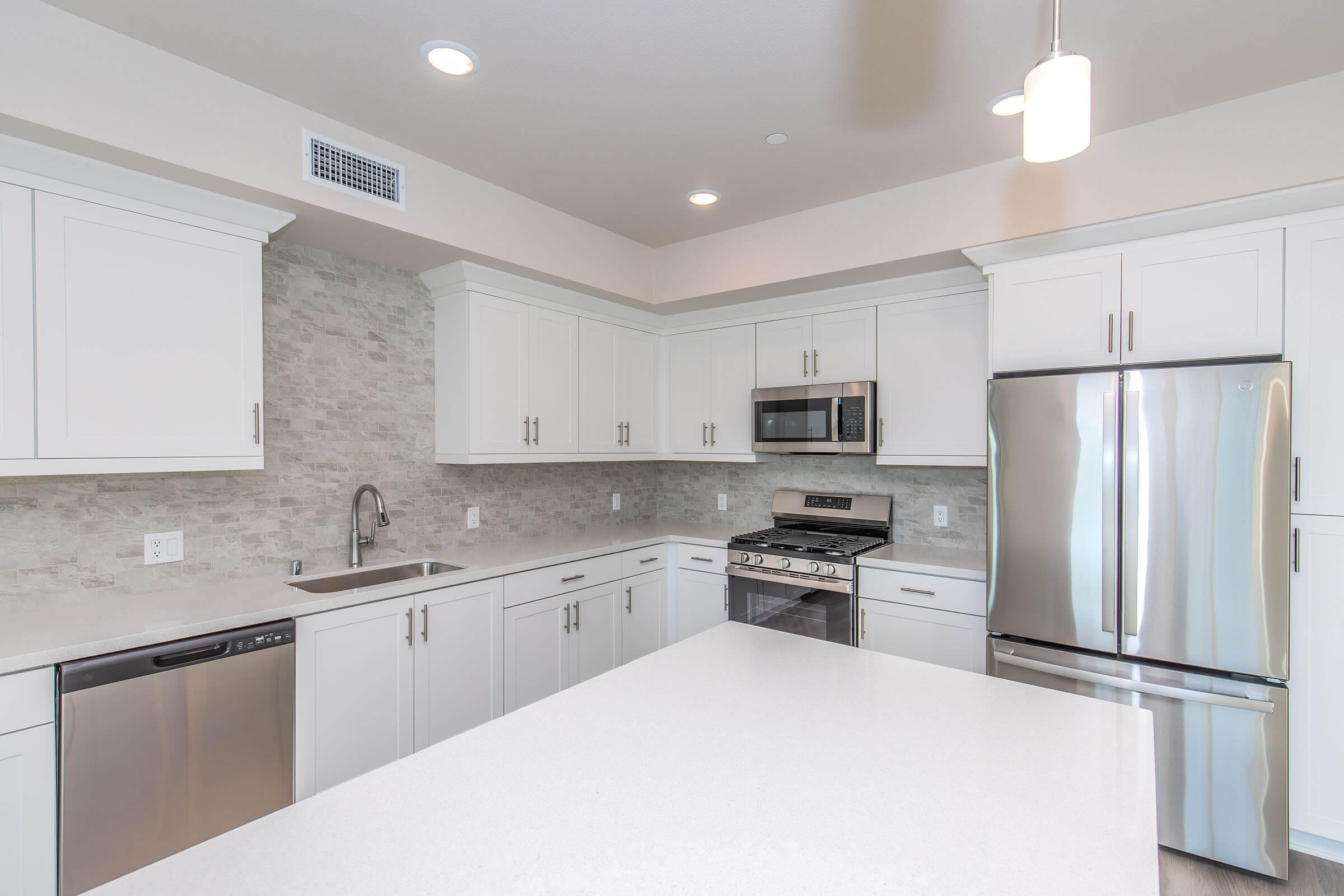 Modern kitchen featuring white cabinets, stainless steel appliances, a large island with a white countertop, and a subtle stone backsplash. The space is well-lit with recessed lighting and has a sleek design, creating a clean and inviting atmosphere.