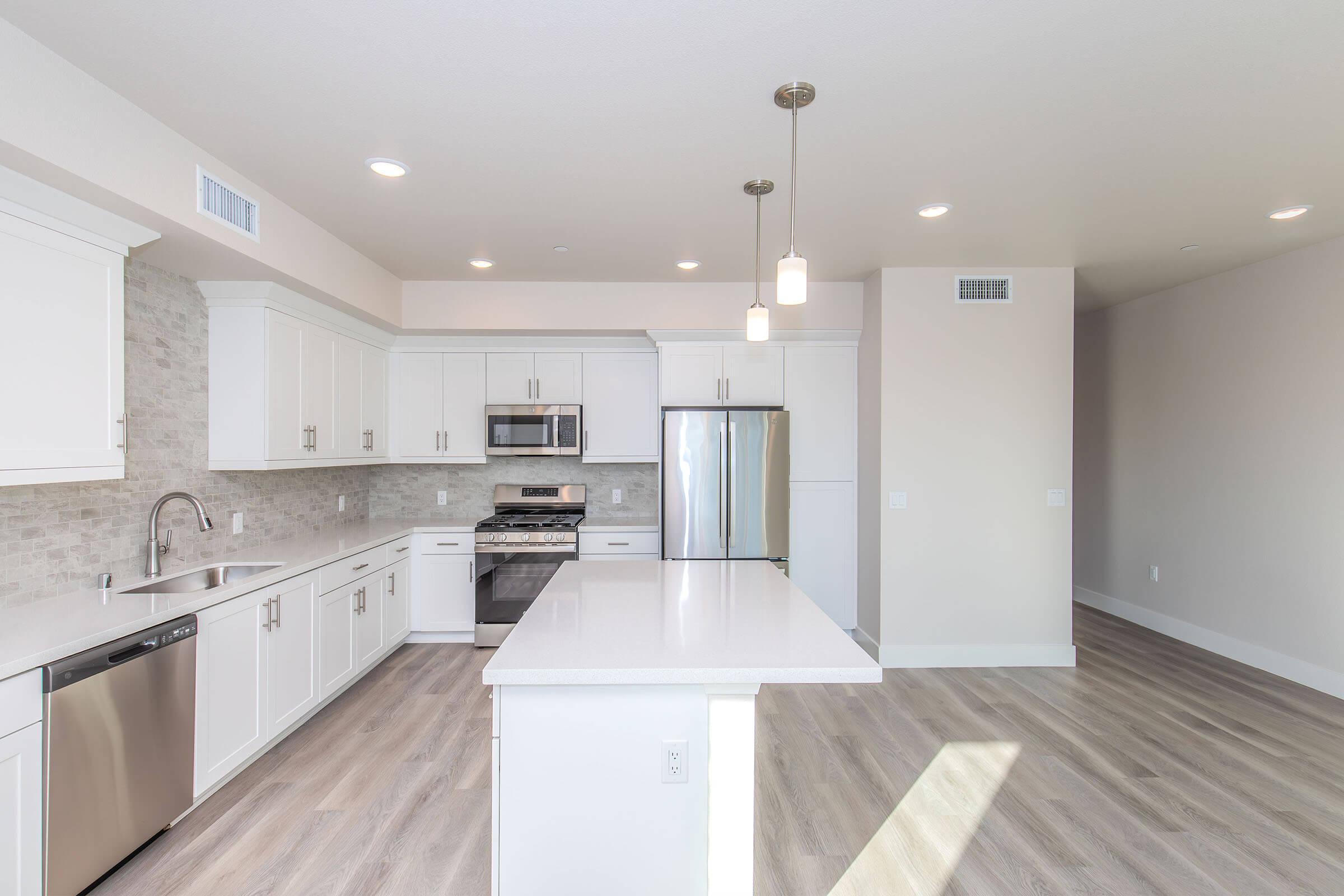 Modern kitchen featuring white cabinetry, stainless steel appliances, and a large island with a countertop. The space is brightly lit with multiple ceiling lights and has light gray flooring. The design is open and airy, creating a contemporary and inviting atmosphere.