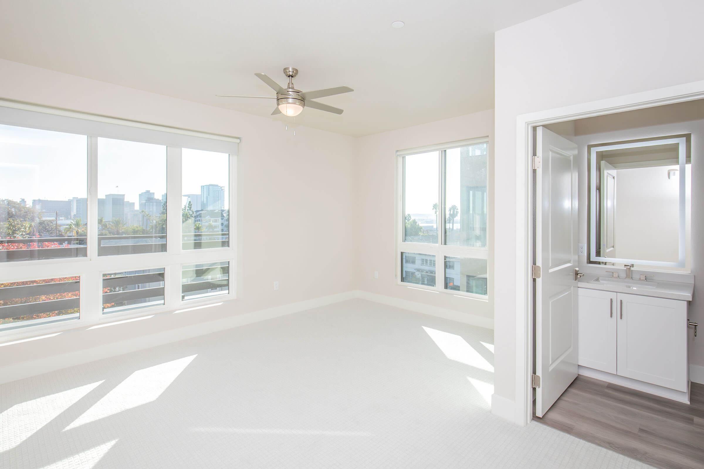 Spacious, bright room with large windows allowing natural light, featuring a ceiling fan. A closed bathroom door is visible, and the space has light-colored walls and flooring, creating a clean and airy atmosphere.