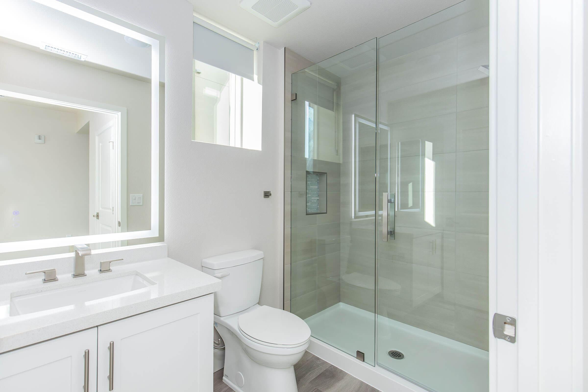 A modern bathroom featuring a glass shower stall, white cabinetry, a sleek countertop with sink, and a toilet. Natural light comes through a window, highlighting the neutral color scheme and clean lines of the design.