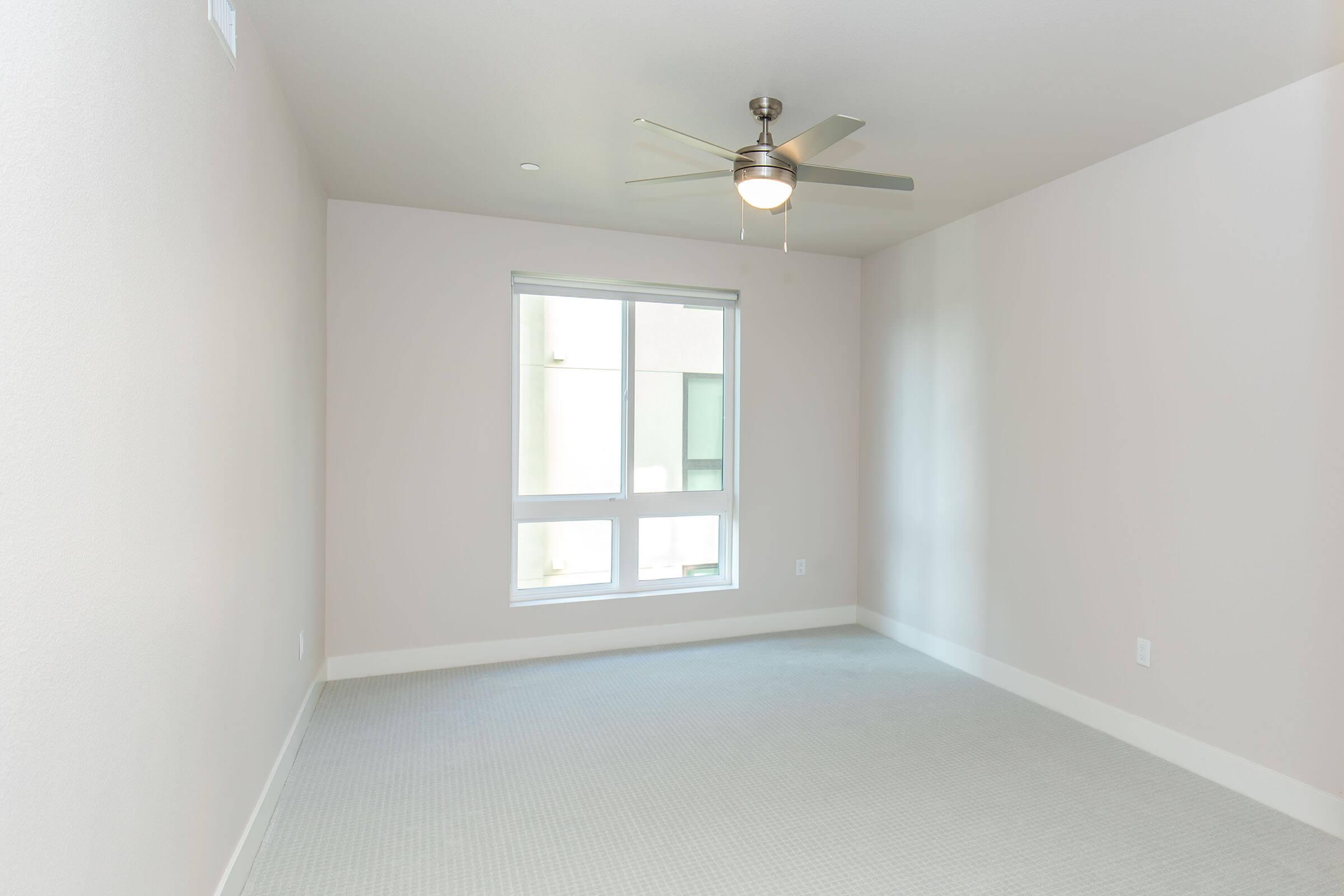 A vacant room with light-colored walls and a window allowing natural light. There is a ceiling fan in the center of the room, and the floor is covered with a light gray carpet. The overall atmosphere is clean and simple, making it suitable for personal customization.