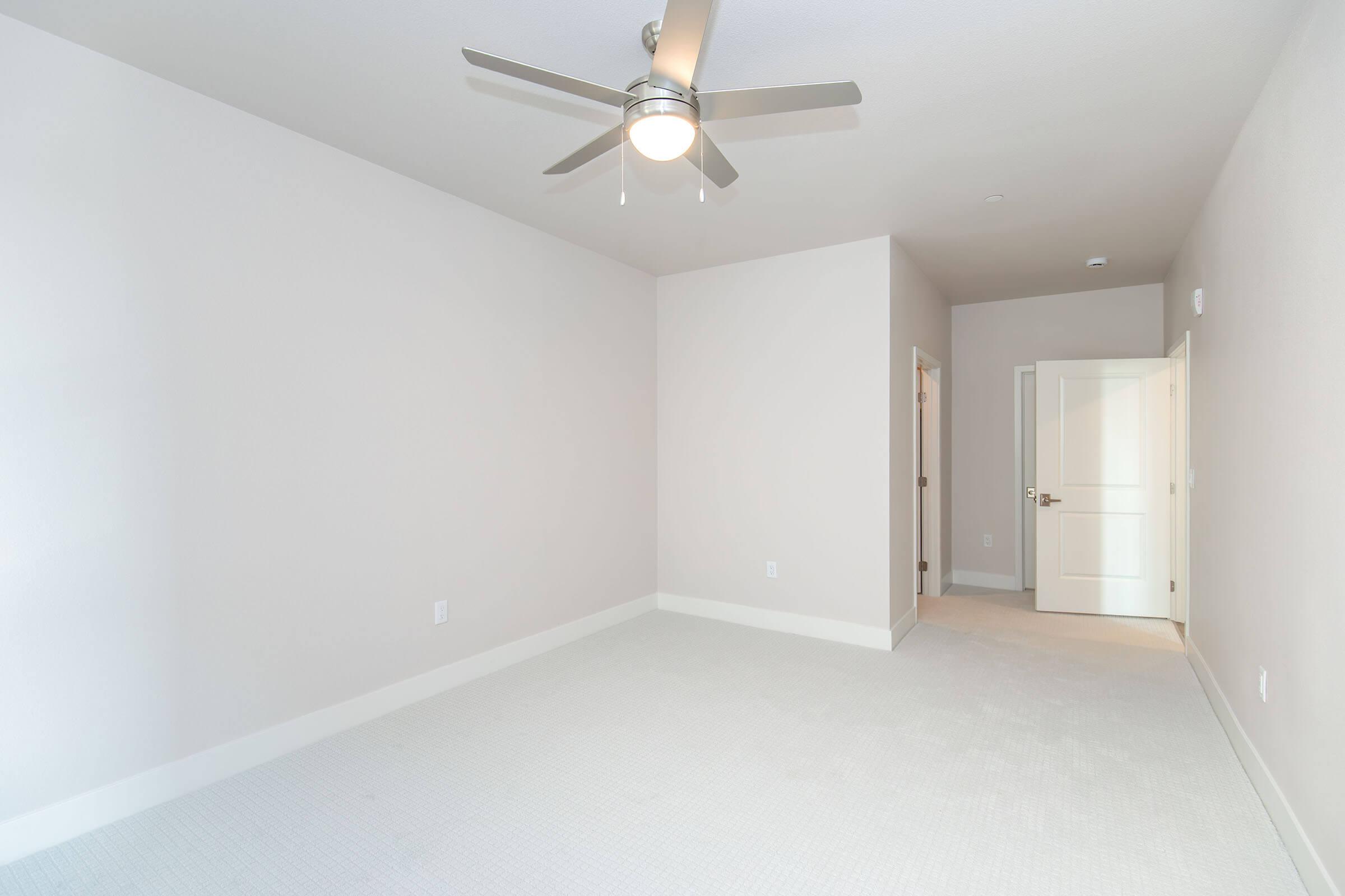 A spacious, empty bedroom with light beige walls and a soft carpeted floor. A ceiling fan is installed, and there is a closed door on one side and an open door leading to another room. Natural light illuminates the space, creating a clean and fresh atmosphere.