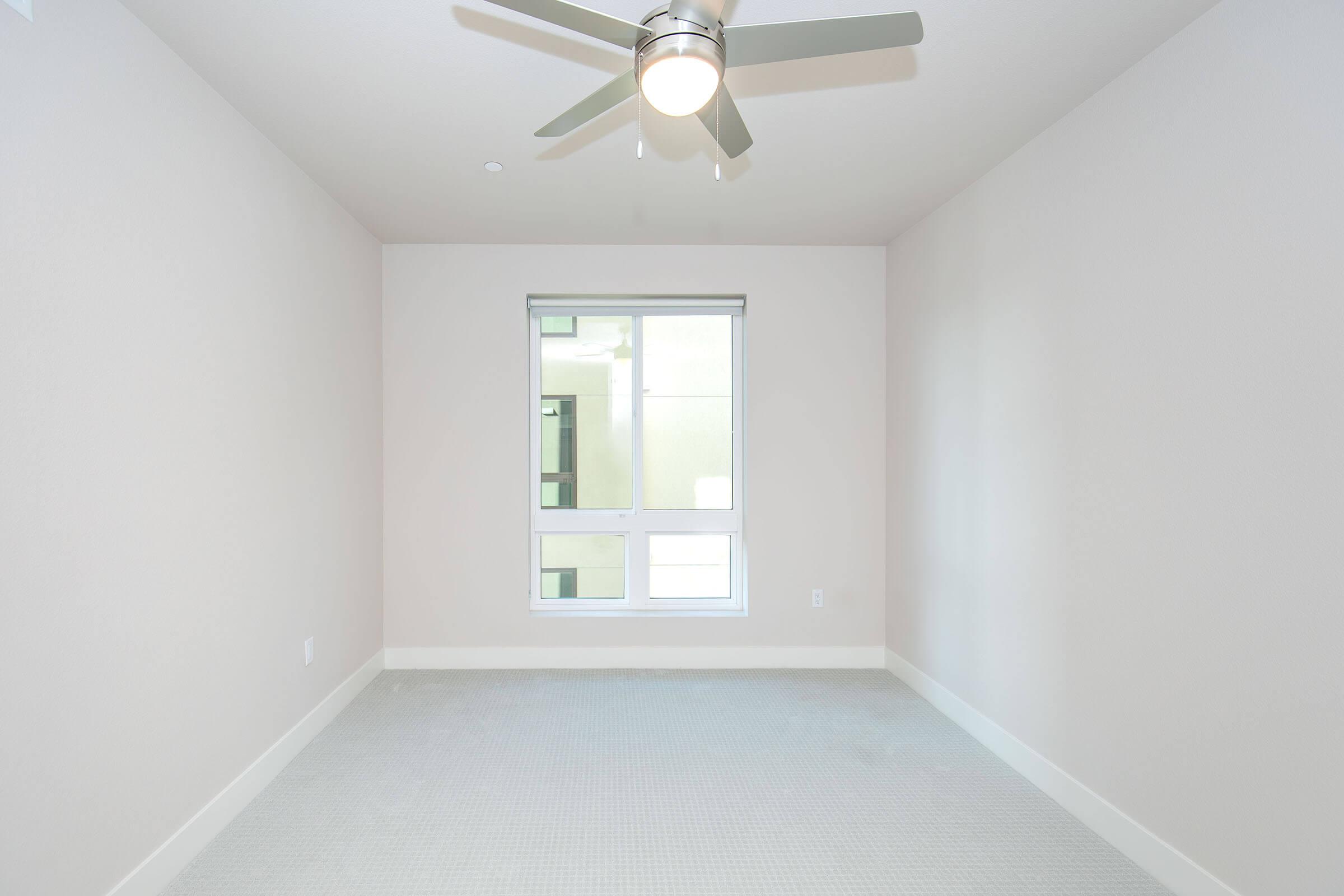 Empty room with light gray walls and a ceiling fan. A large window lets in natural light, facing another structure. The floor is carpeted in a light color, creating a simple and minimalist atmosphere.