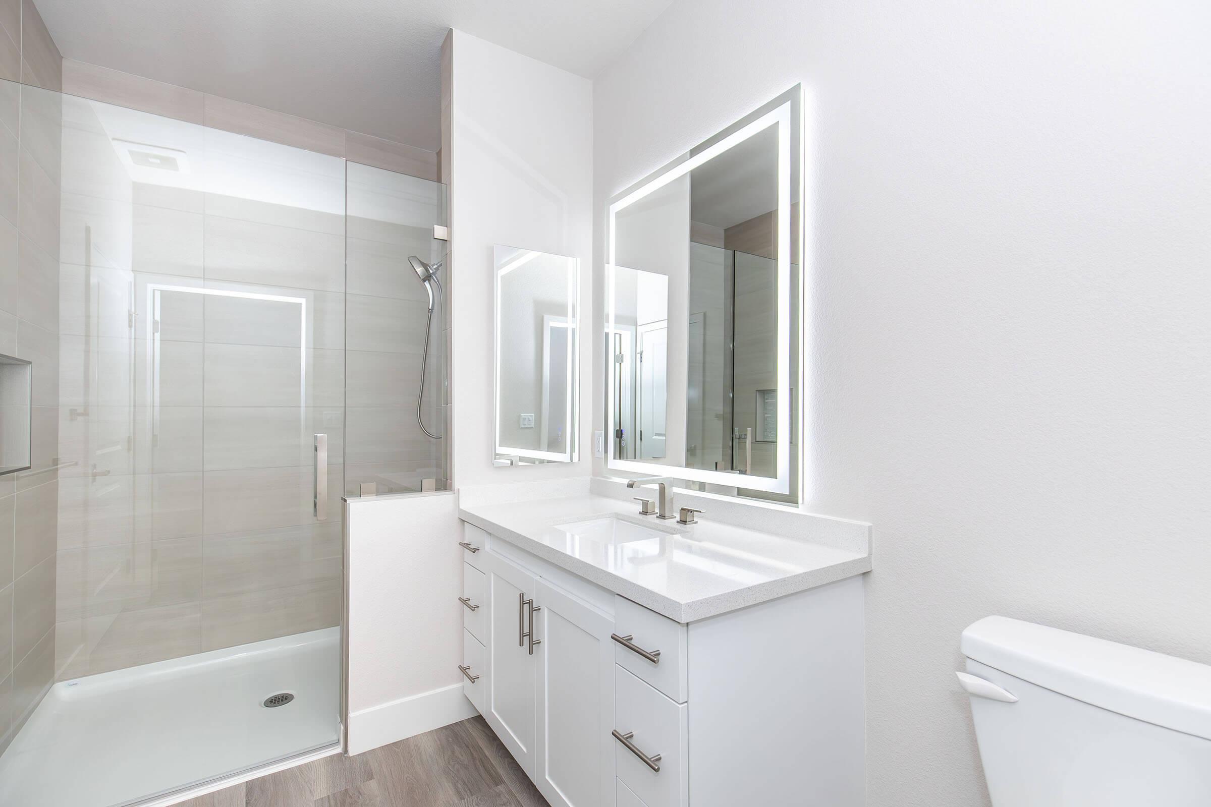 Modern bathroom featuring a sleek design with a glass shower enclosure, double vanity with under-mounted sinks, backlit mirrors, and light-colored tile walls. The toilet is positioned to the right, and the flooring is a light wood-like material, creating a clean and spacious look.