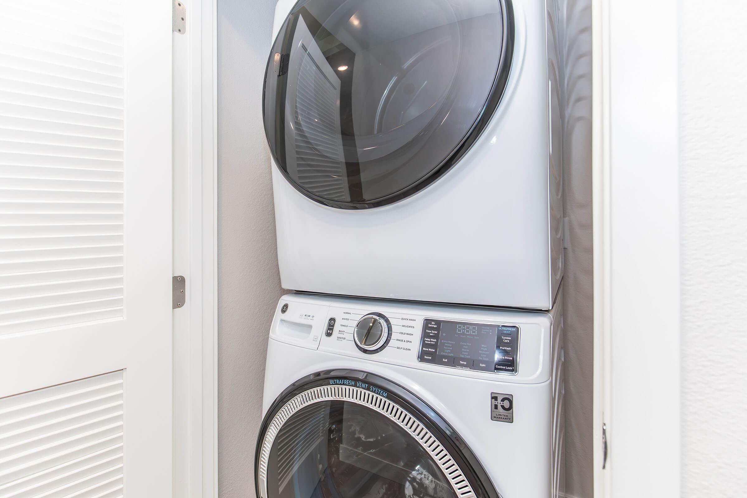 A stacked washer and dryer set in a small laundry space, featuring a modern design with a white finish. The washer is on the bottom, and the dryer is on top, with a front-loading door. The appliances are positioned next to closed bifold doors and a light-colored wall.