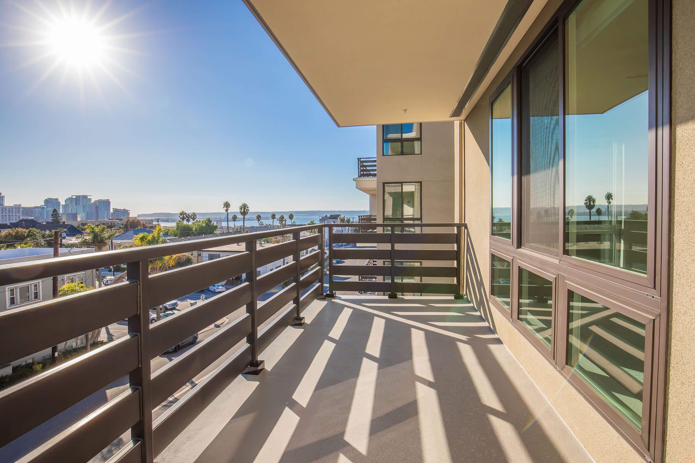 Spacious balcony with a modern railing, overlooking a vibrant cityscape and waterfront. Bright sunlight casts shadows on the floor, with palm trees visible in the foreground and buildings in the background, creating a serene and inviting outdoor space.