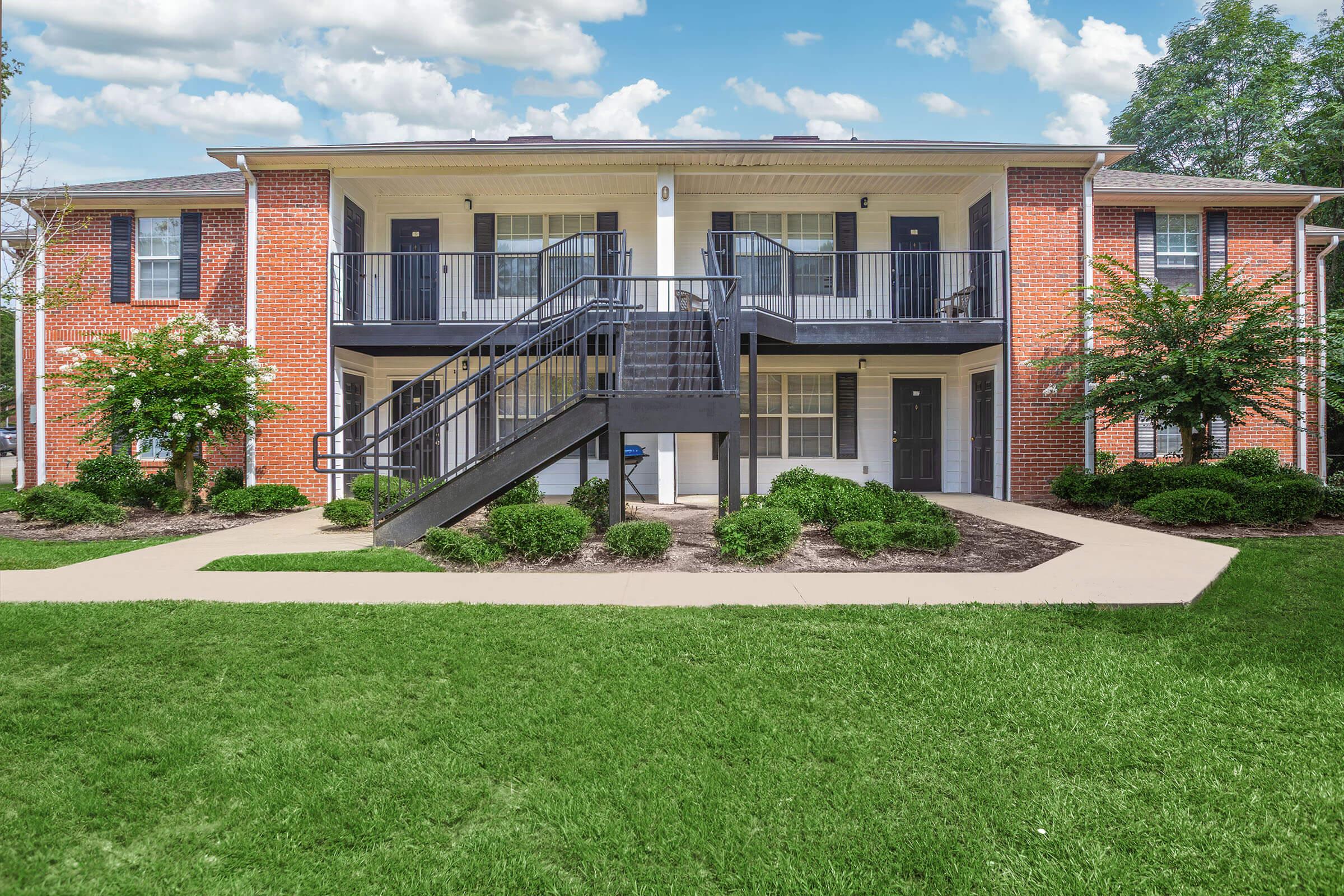 Two-story brick apartment building featuring balconies, outdoor staircases, and well-maintained landscaping. Lush green grass in the foreground with freshly trimmed shrubs and trees surrounding the building. The sky is partly cloudy, adding a bright atmosphere to the scene.