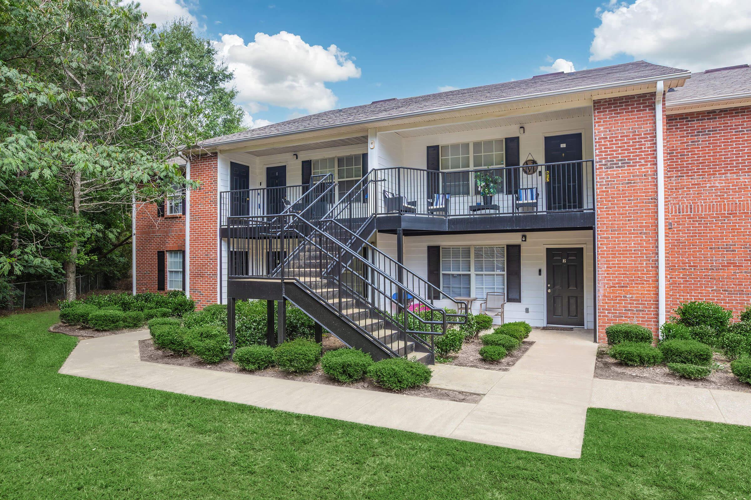 Two-story apartment building with a brick exterior and black metal stairway leading to the upper level. The ground floor features two sets of doors and windows, surrounded by neatly trimmed shrubs and a green lawn. The sky is clear with a few clouds.