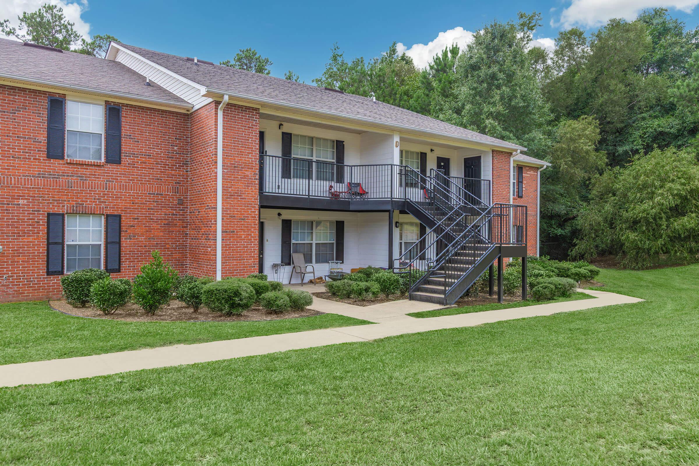 Two-story brick apartment building with black railings and covered balconies. The front yard features manicured grass and shrubs, with a concrete pathway leading to the entrance. The background includes trees and a clear blue sky with a few clouds.