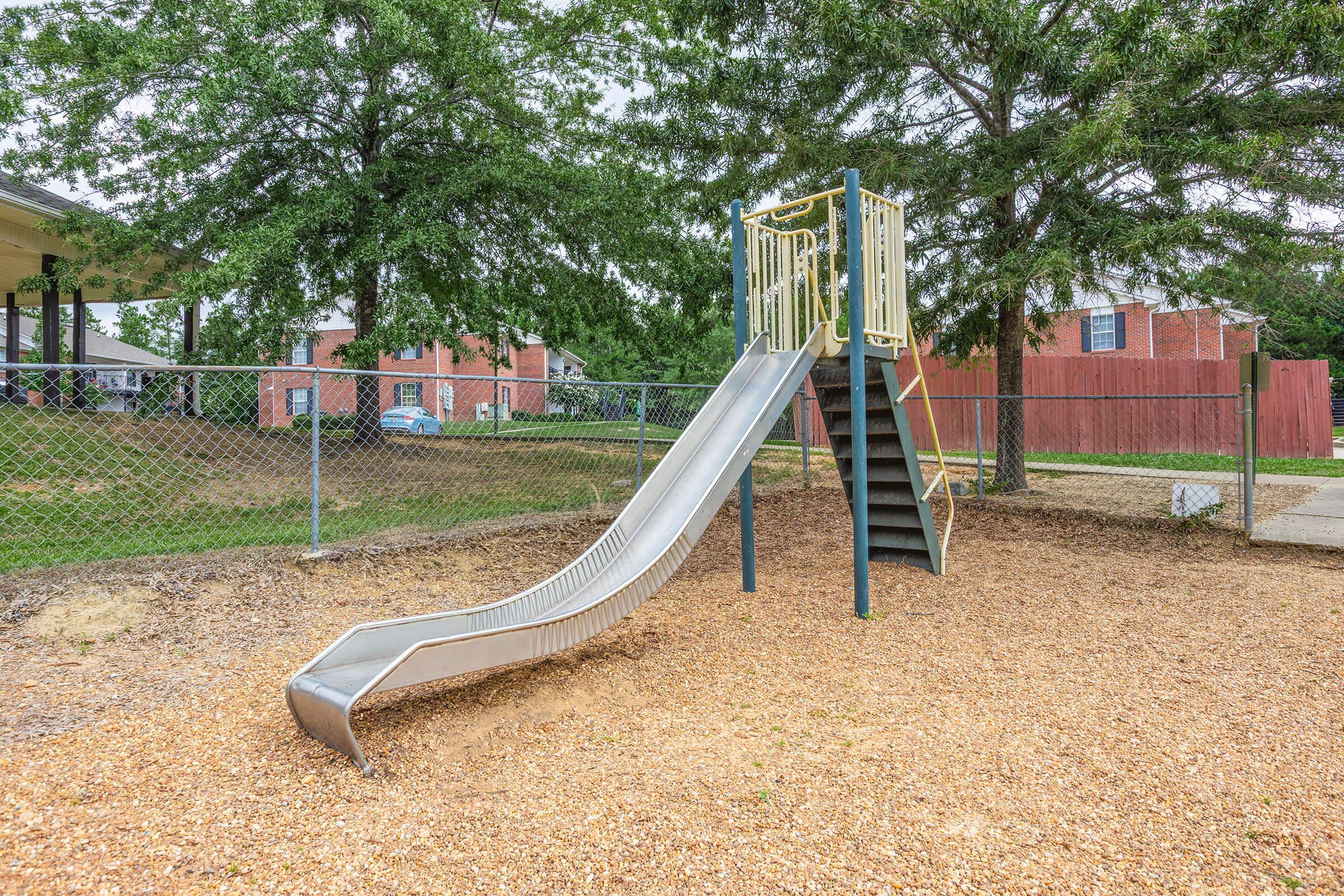 A playground slide made of metal stands beside a fenced area, surrounded by trees. The slide has a small set of stairs leading up to it and is situated on a gravel surface. In the background, there are residential buildings partially visible through the trees.