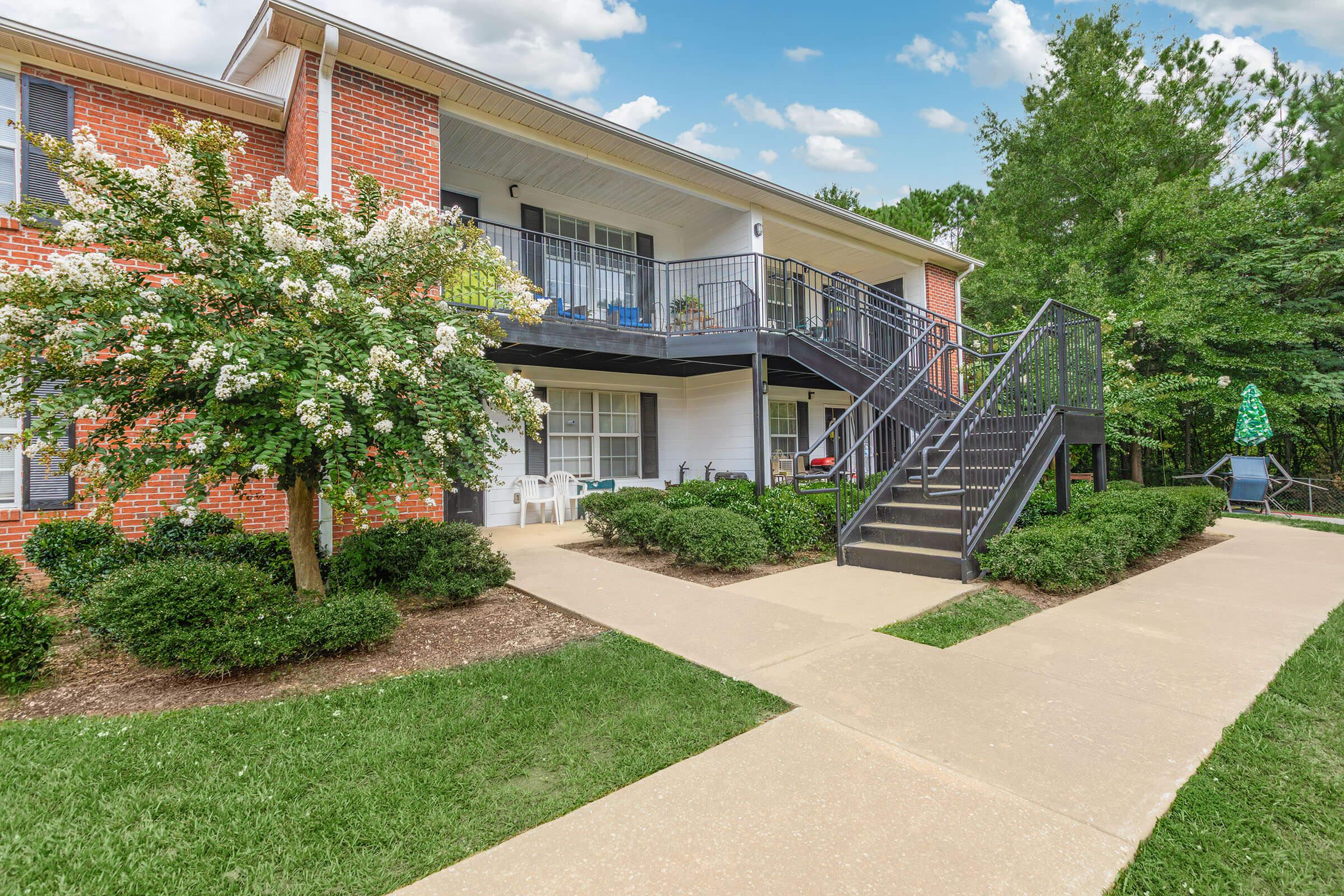 A well-maintained two-story apartment building featuring brick and white siding. The entrance has a staircase leading to an upper balcony, surrounded by vibrant green landscaping, shrubs, and a flower tree. Sunlight brightens the scene, highlighting the inviting path to the entrance.